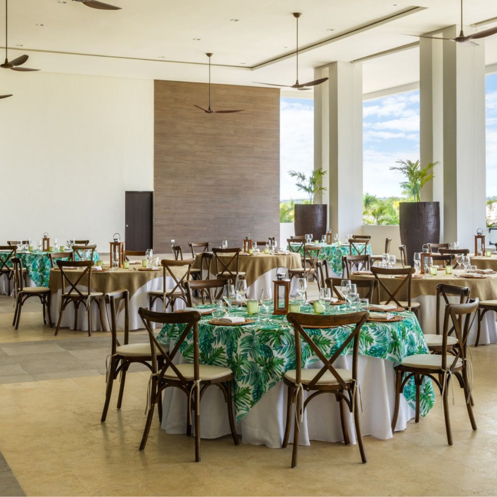 A wedding dining hall adorned with round tables draped in green leaf-patterned tablecloths and complemented by wooden chairs creates the perfect setting for a destination wedding.