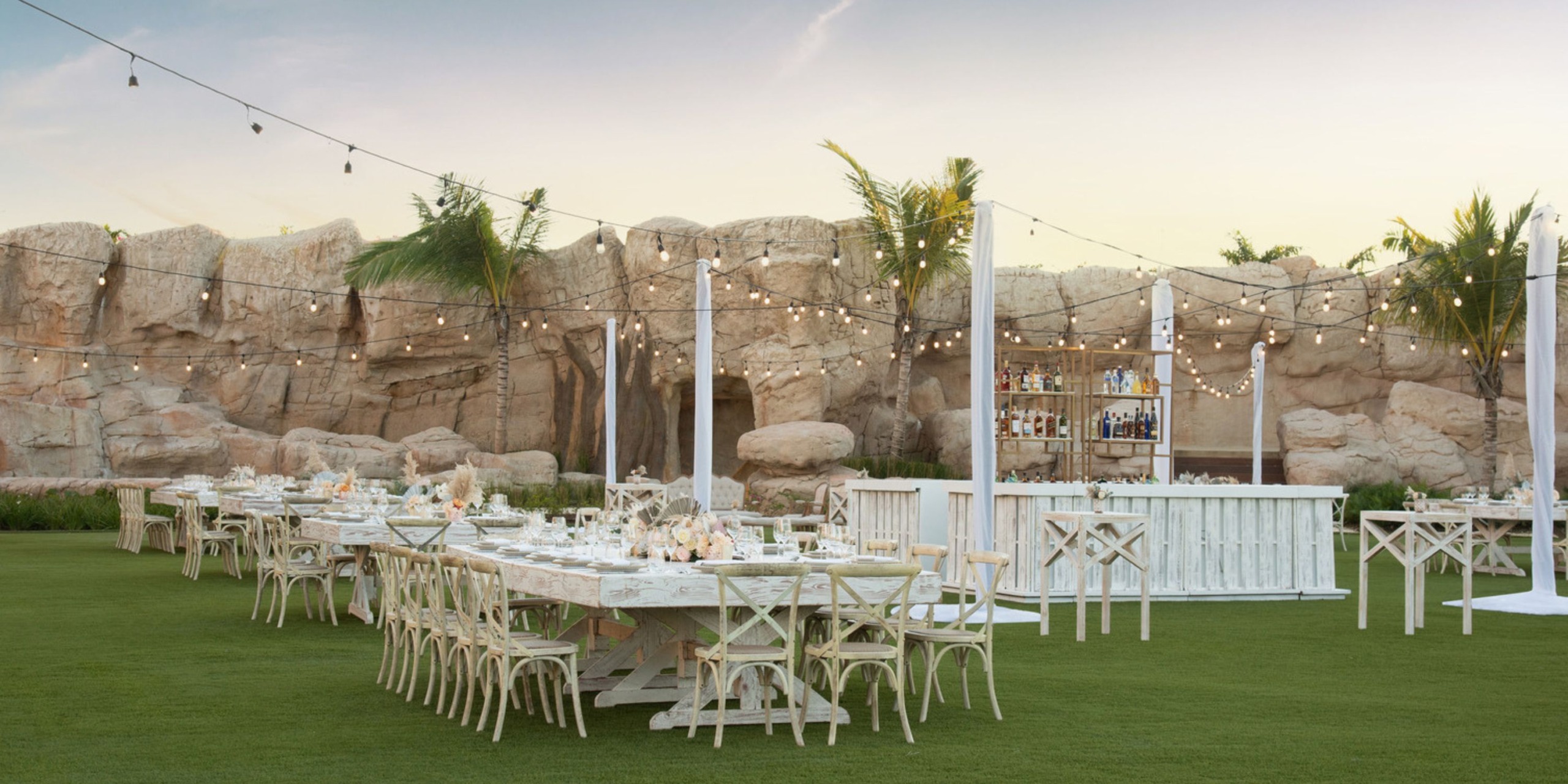 A charming outdoor dining setup for a destination wedding, featuring string lights, wooden tables, and chairs arranged on a lush grassy area.