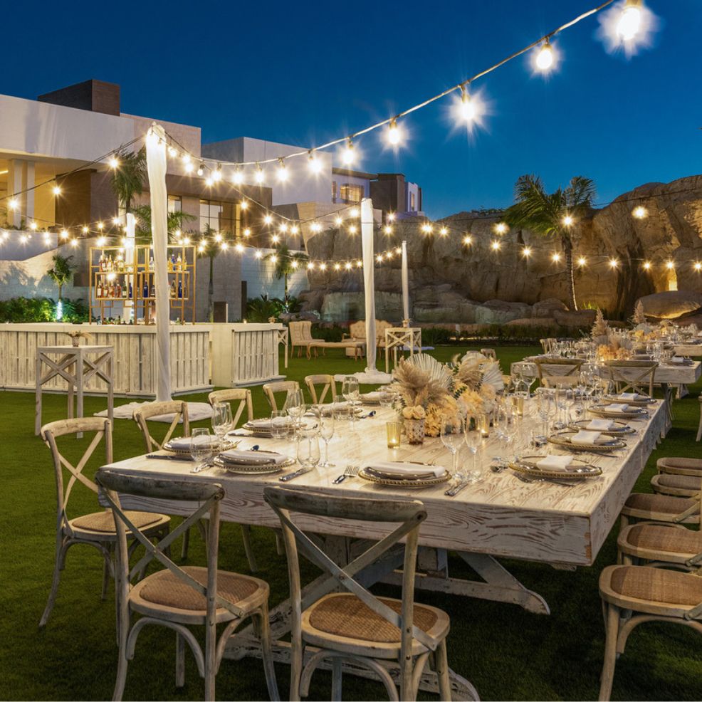 A picturesque destination wedding scene featuring an outdoor dining area adorned with string lights, elegantly arranged tables, and chairs nestled in a nighttime garden ambiance.