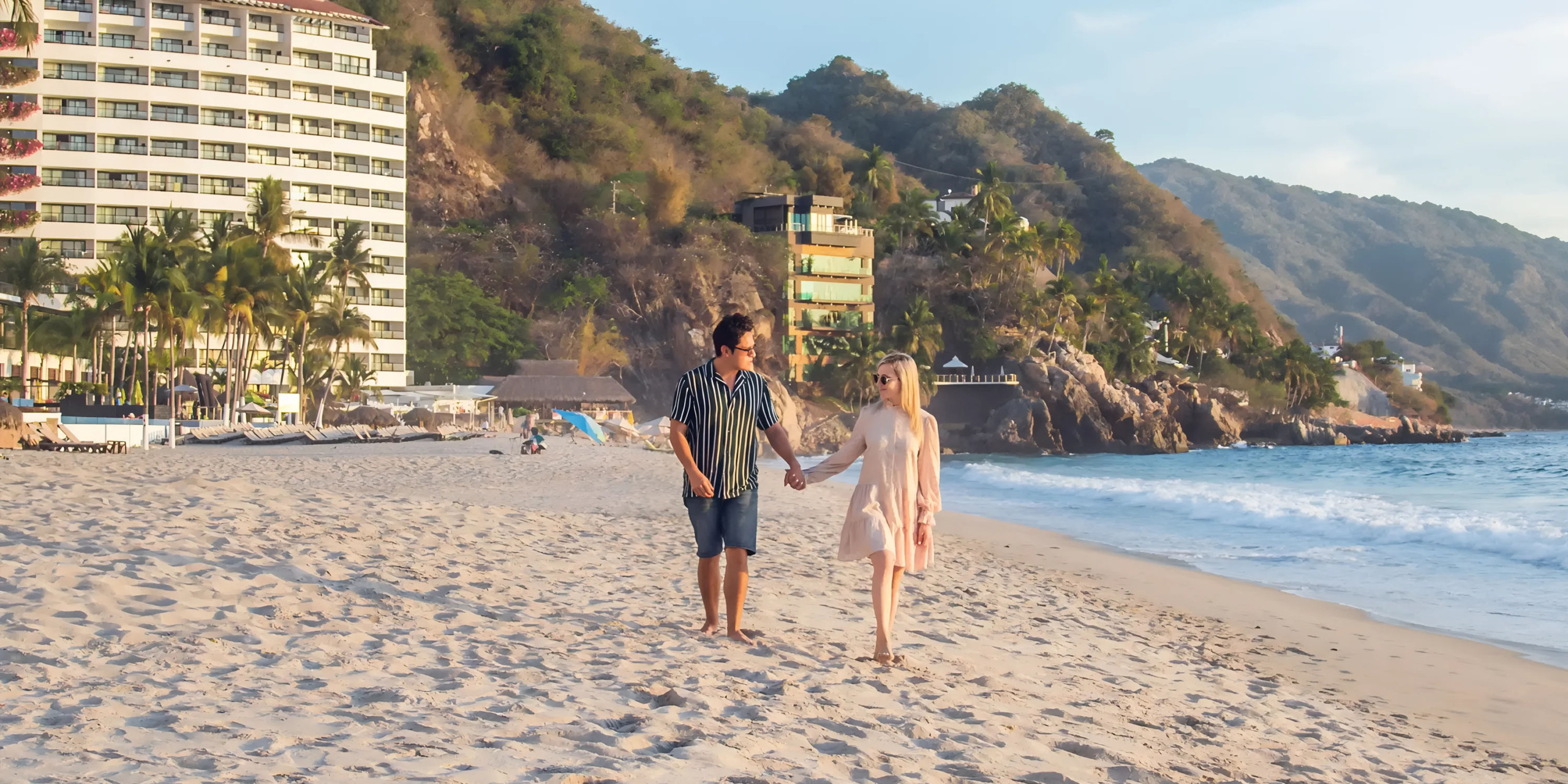 Couple holding hands and walking along a sandy beach with buildings in the background during sunset at Hyatt Ziva Puerto Vallarta.