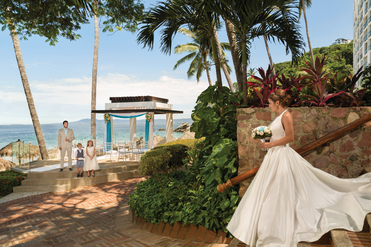 The bride makes her way towards the groom and children by a tropical altar at the Hyatt Ziva Puerto Vallarta beach, creating a stunning setting for their destination wedding.