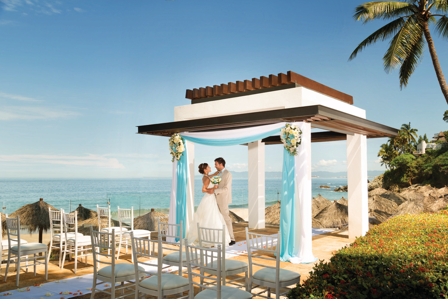 The bride and groom are celebrating their destination wedding under a beautifully decorated gazebo at Hyatt Ziva Puerto Vallarta beach, surrounded by elegantly arranged chairs.