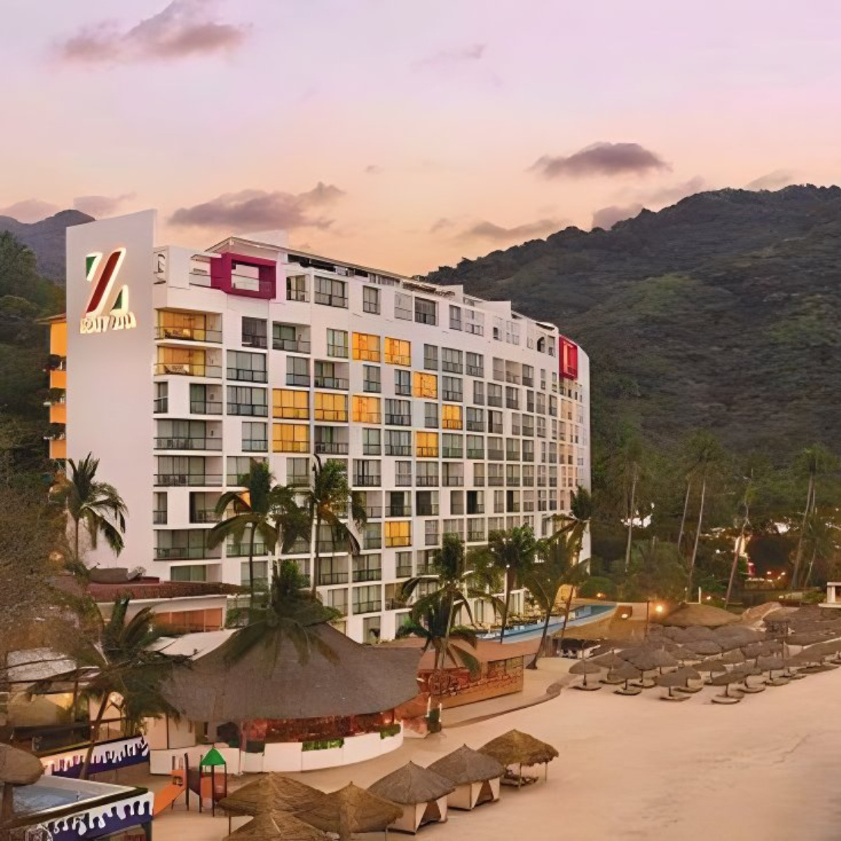 A Hyatt Ziva Puerto Vallarta by a beach at sunset, ideal for a destination wedding, featuring a lit-up facade, palm trees, poolside loungers, and mountain backdrop.