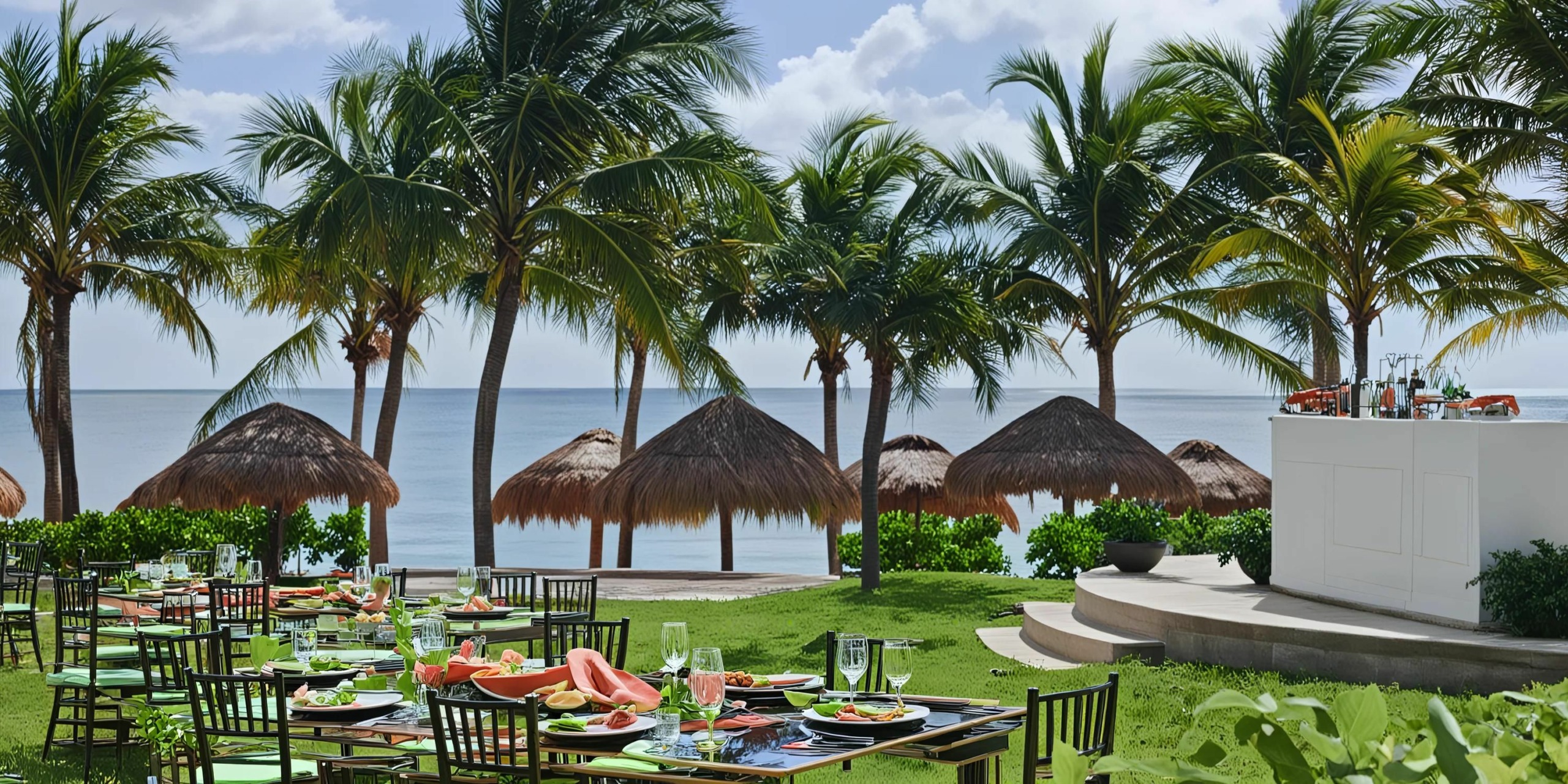 Enjoy an outdoor dining setup at the Hyatt Ziva Riviera Cancun, featuring tables and chairs arranged on lush grass with a stunning ocean view, surrounded by graceful palm trees and charming thatched-roof structures.