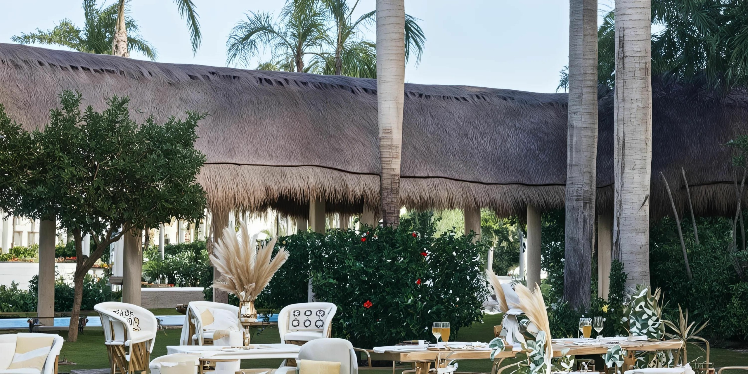 An outdoor seating area with white chairs and tables is set in a lush garden at the Hyatt Ziva Riviera Cancun, with palm trees and thatched-roof structures visible in the background.