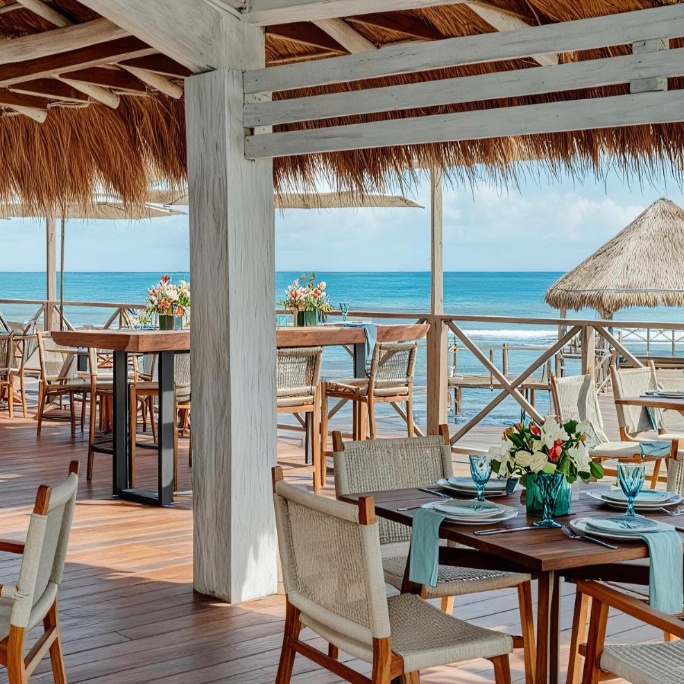 An open-air dining area with wooden tables and chairs is set on a deck with a thatched roof, overlooking the ocean.