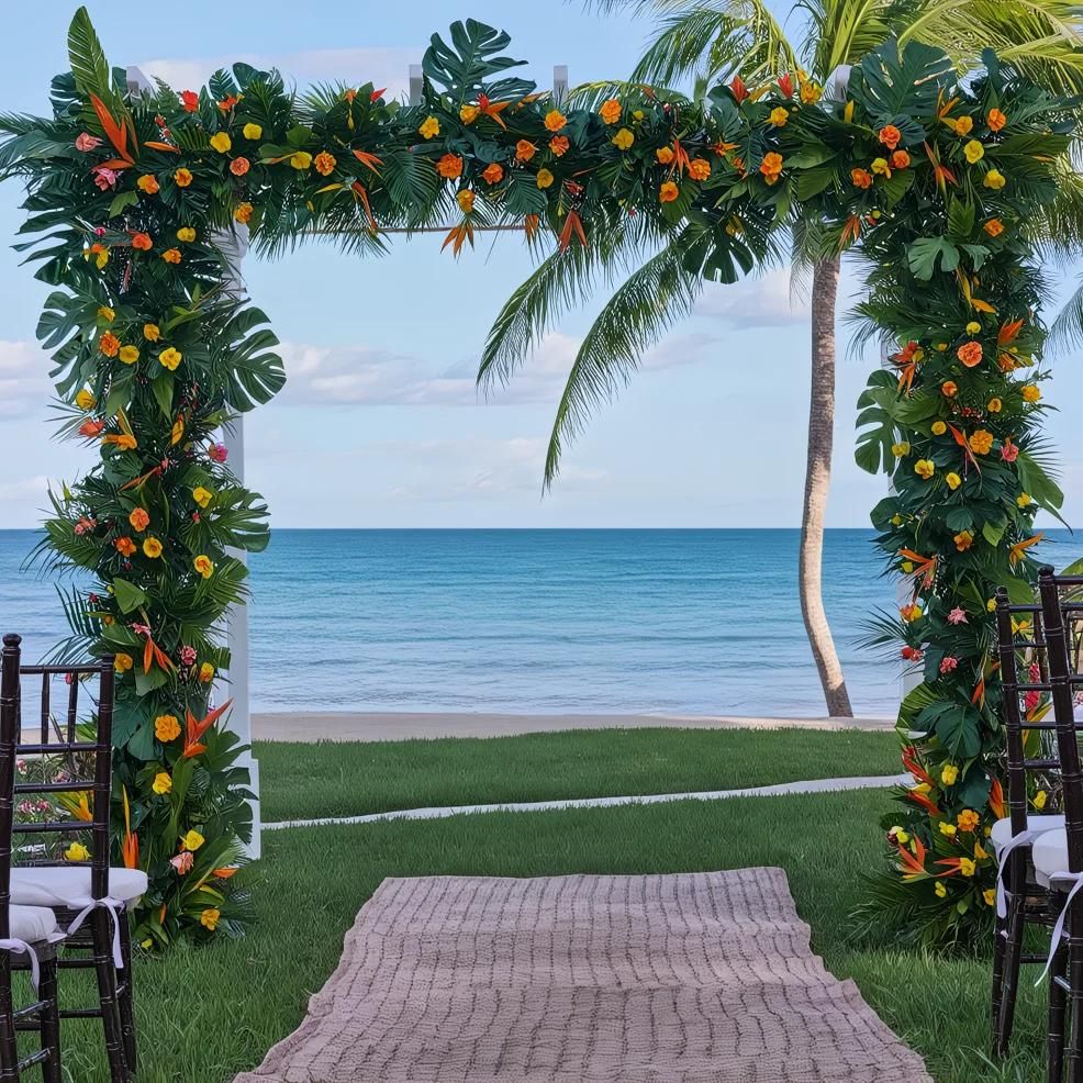 An outdoor wedding arch decorated with tropical flowers overlooks a beach with clear blue water and a single palm tree. Chairs are arranged on either side of a rug aisle leading to the arch.
