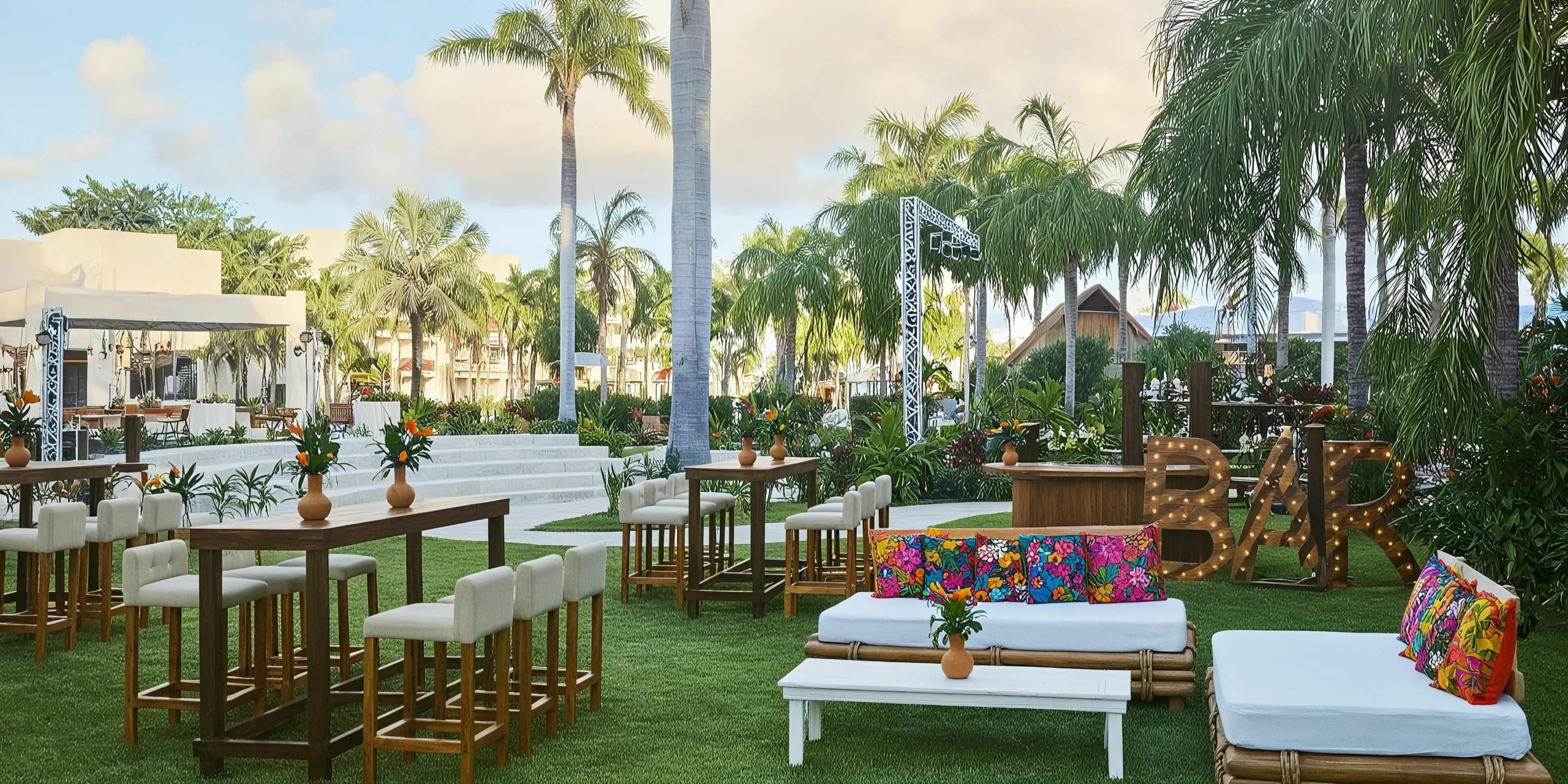 The outdoor event space at Hyatt Ziva Riviera Cancun features lush green grass, elegant palm trees, wooden tables paired with white chairs, and cozy sofas adorned with colorful cushions. In the background, a stylish bar area highlighted by a decorative "BAR" sign adds to the vibrant atmosphere.