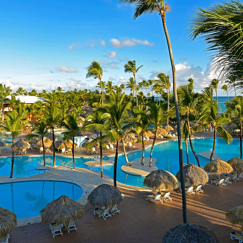 Iberostar Dominicana, a destination wedding venue with a large swimming pool surrounded by palm trees and thatched umbrellas, under a clear blue sky.