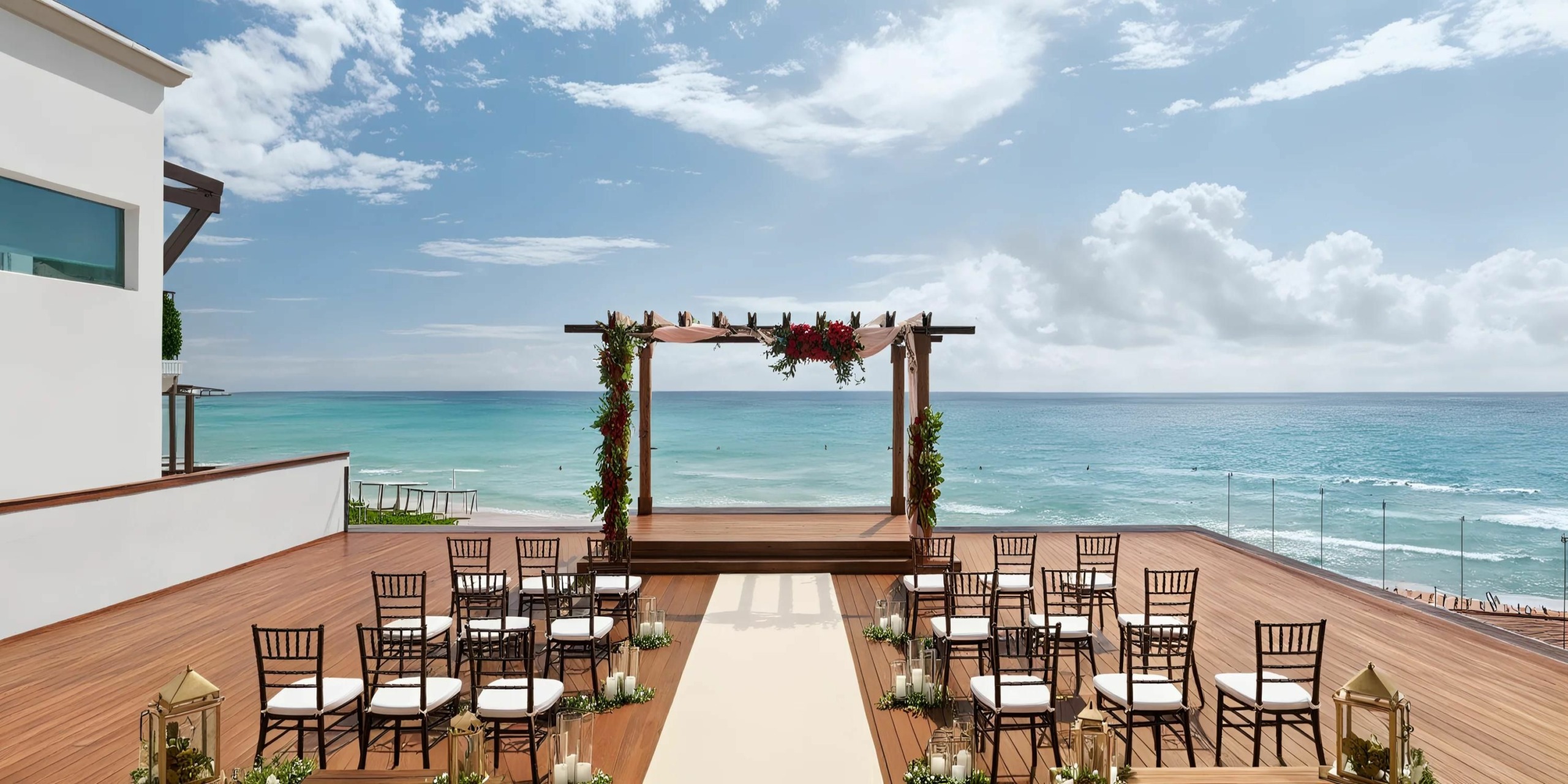 Hilton Playa del Carmen offers an outdoor beachside wedding setup featuring a wooden deck with chairs arranged to face a floral-decorated archway, complemented by the serene backdrop of the ocean and sky.