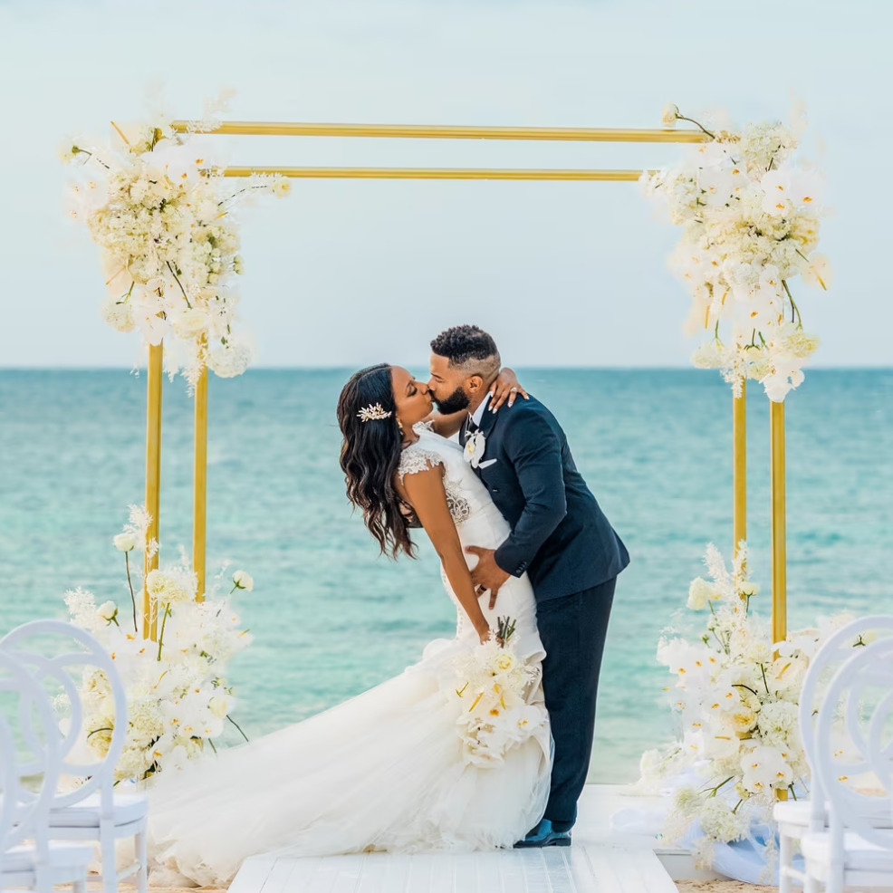 A couple kisses under a floral arch at a destination wedding ceremony at Iberostar Selection Rose Hall, with the ocean in the background.