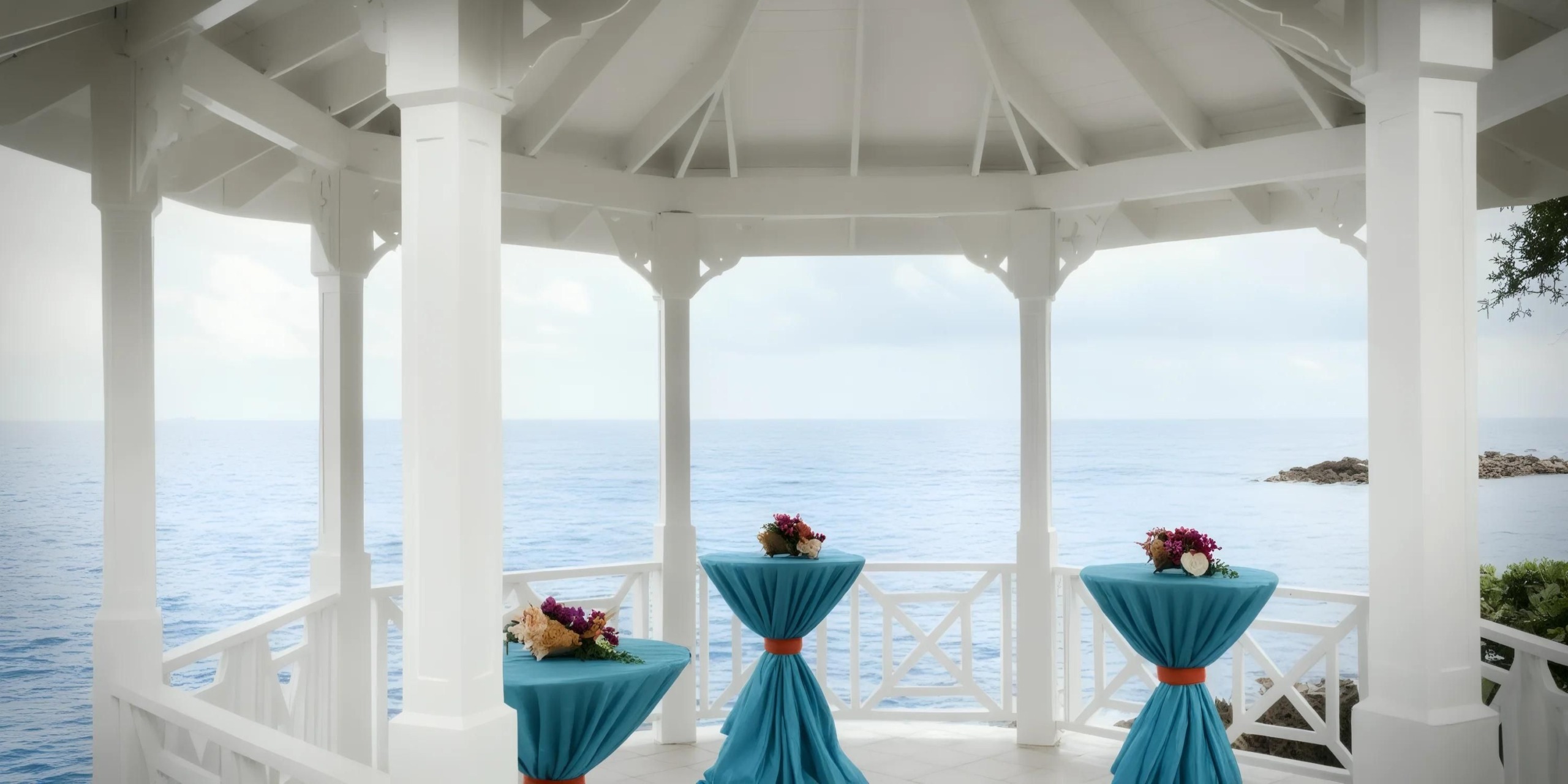 White gazebo overlooking the ocean, with three tall tables covered in blue cloth and floral centerpieces.