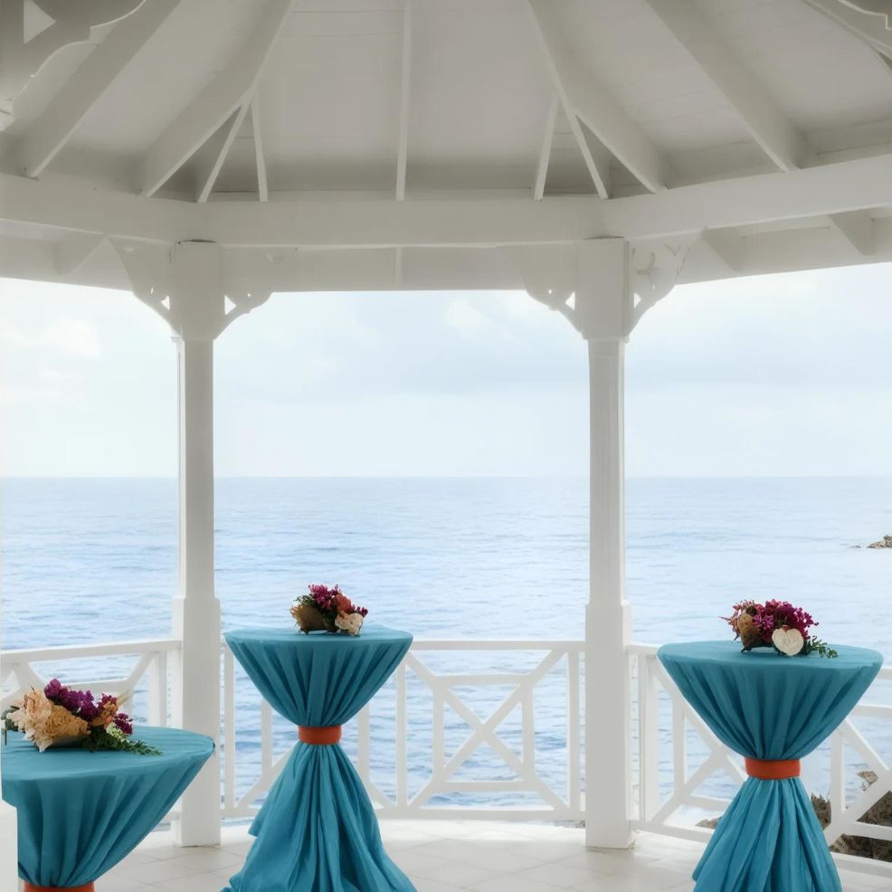 A gazebo with a view of the ocean, featuring three tall tables covered in turquoise cloth and topped with flower arrangements.
