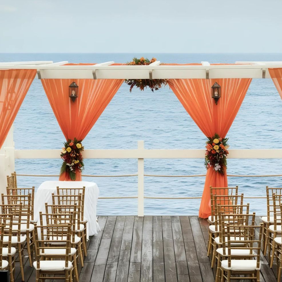 Outdoor wedding setup with orange drapes and floral arrangements overlooking the ocean. Rows of wooden chairs face a small table under a pergola-style structure on a wooden deck.
