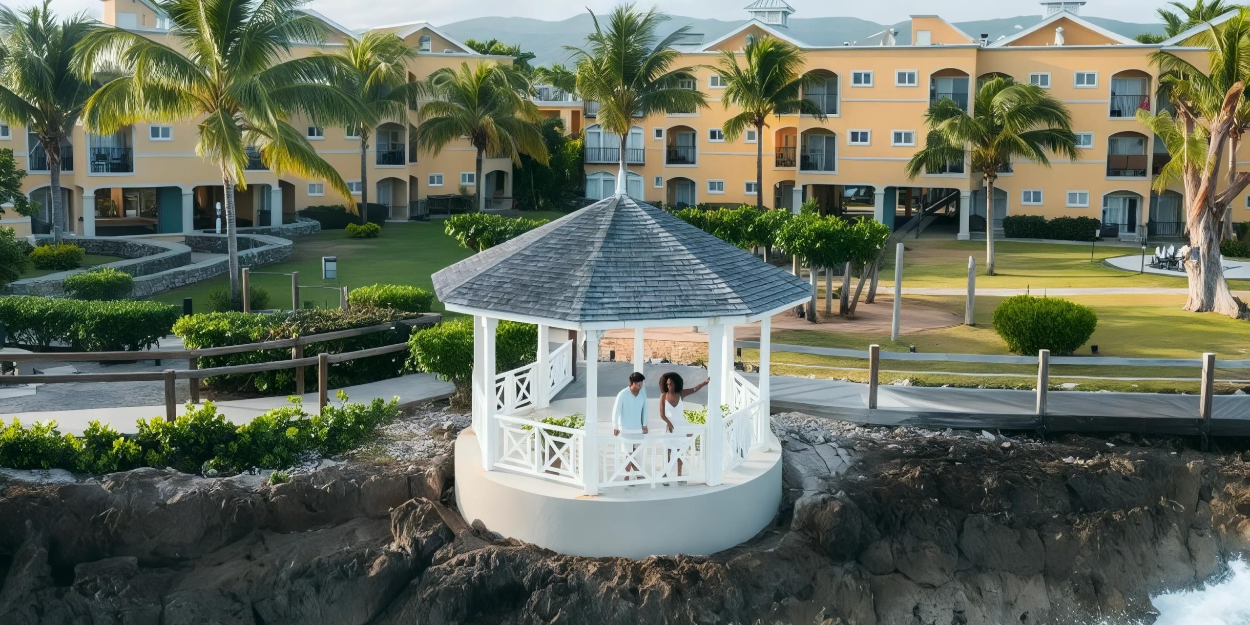 A white gazebo overlooks a rocky shore with people inside, surrounded by palm trees and facing a yellow resort building.