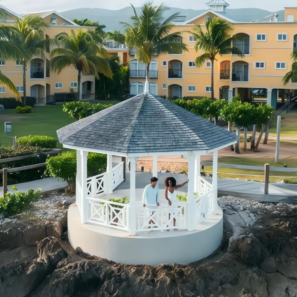 A couple stands in a white gazebo overlooking a rocky shore, with palm trees and yellow buildings in the background.