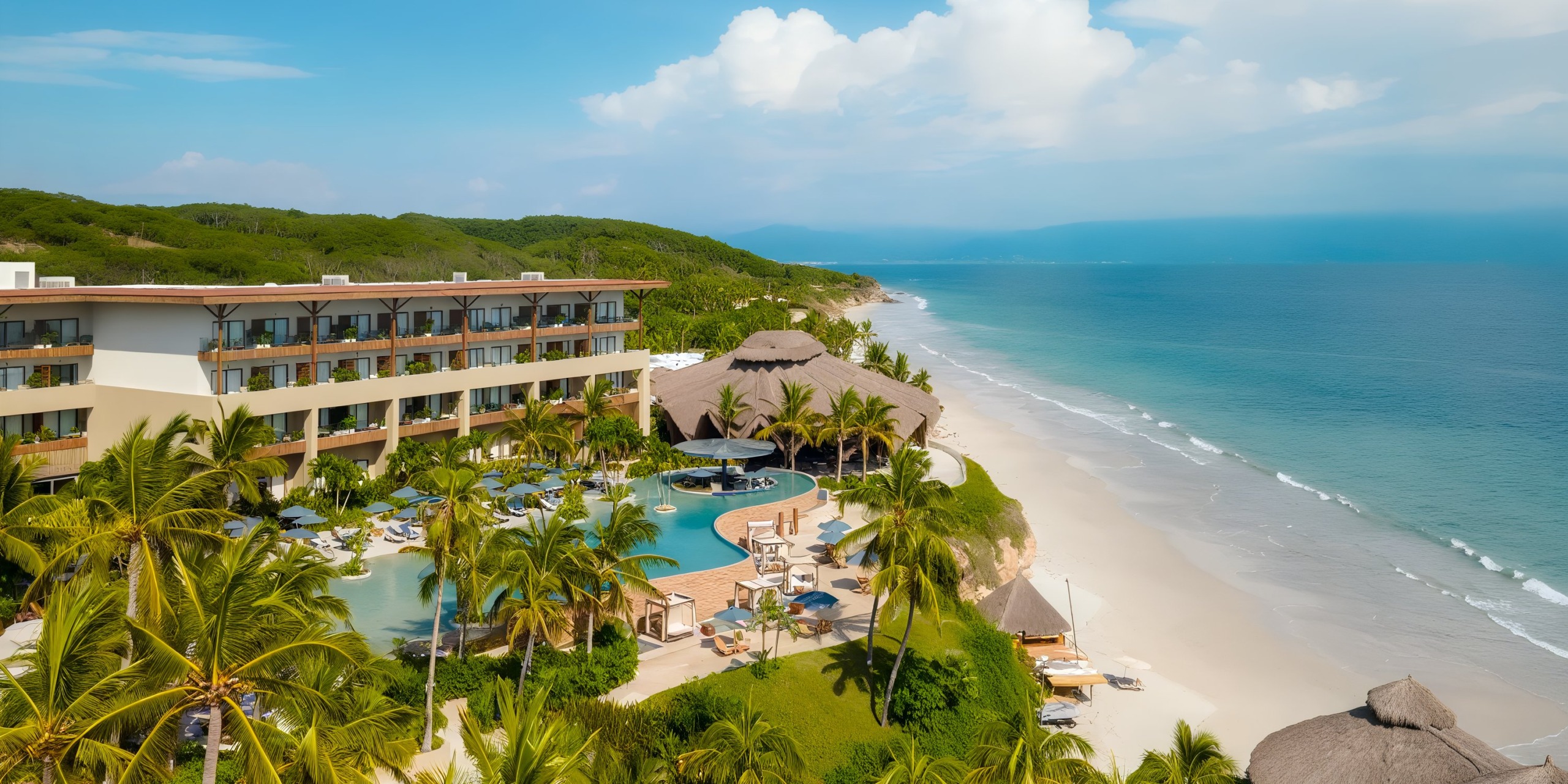 Aerial view of Marival Armony, a beachfront resort featuring a large pool encircled by palm trees, adjacent to a sandy beach and the ocean beneath a partly cloudy sky.