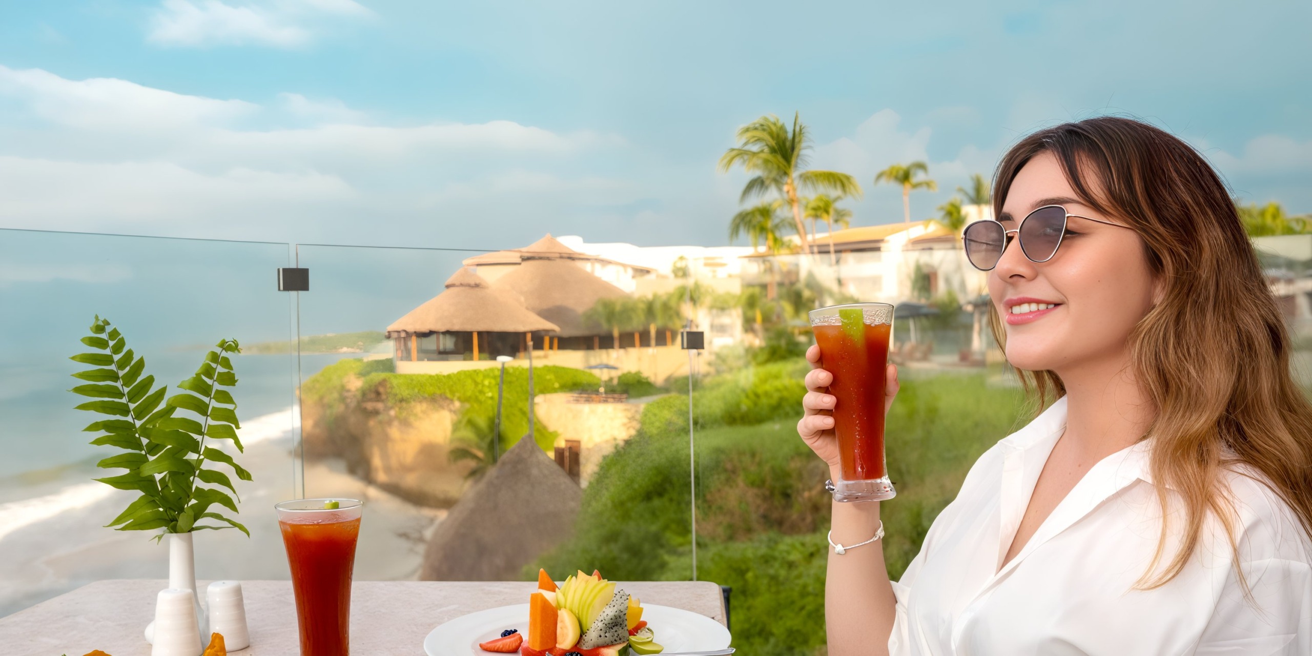 A woman in sunglasses enjoys a drink on the patio of Marival Armony, overlooking a tropical beach adorned with palm trees and thatched-roof buildings.