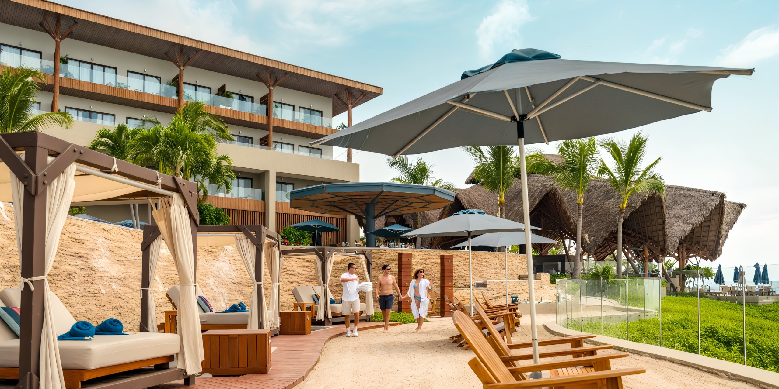 At Marival Armony, enjoy a seaside escape complete with lounge chairs, shaded cabanas, and large umbrellas. Three individuals take a stroll along the path while palm trees and thatched structures provide a picturesque backdrop.