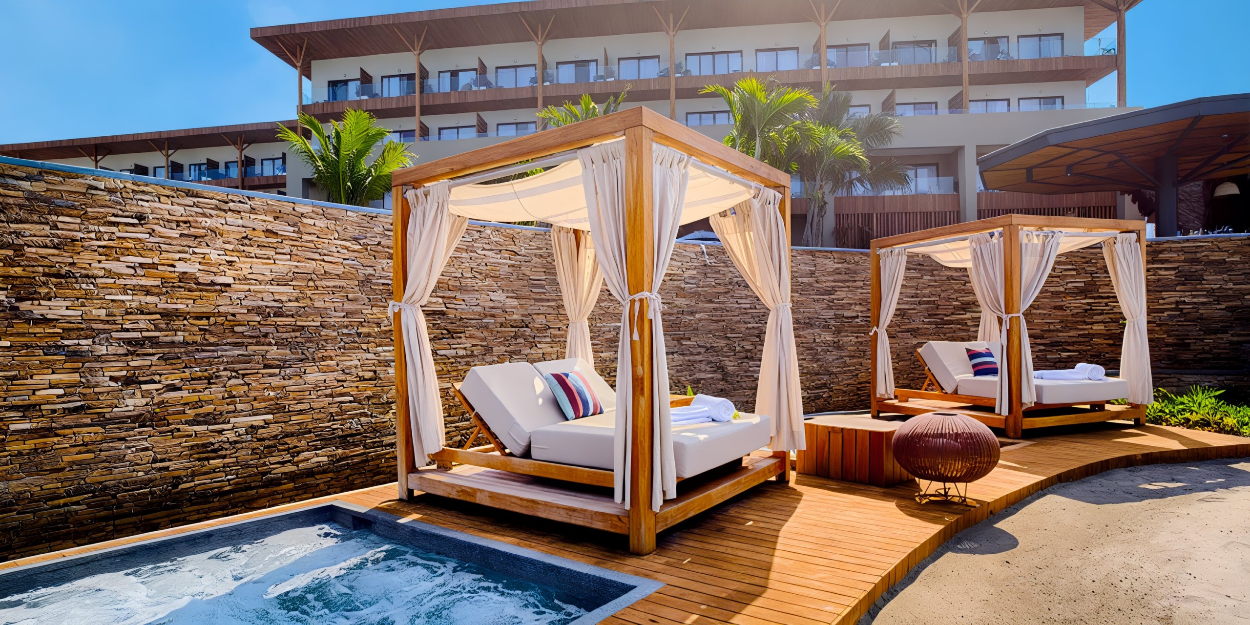 Poolside cabanas featuring white cushions and vibrant pillows are arranged on a wooden deck adjacent to a hot tub, all situated in front of a stone wall and the Marival Armony resort building.