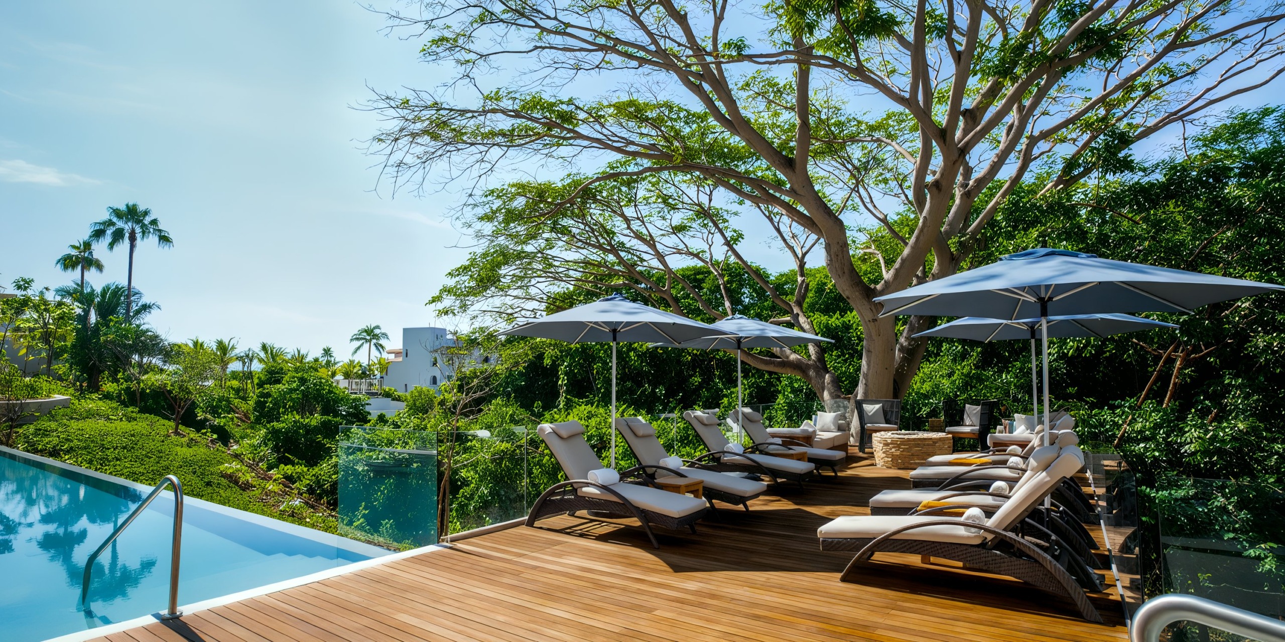 The Marival Armony features a wooden deck complete with lounge chairs and umbrellas, which offers a stunning view of an infinity pool surrounded by lush greenery and towering trees beneath a clear blue sky.