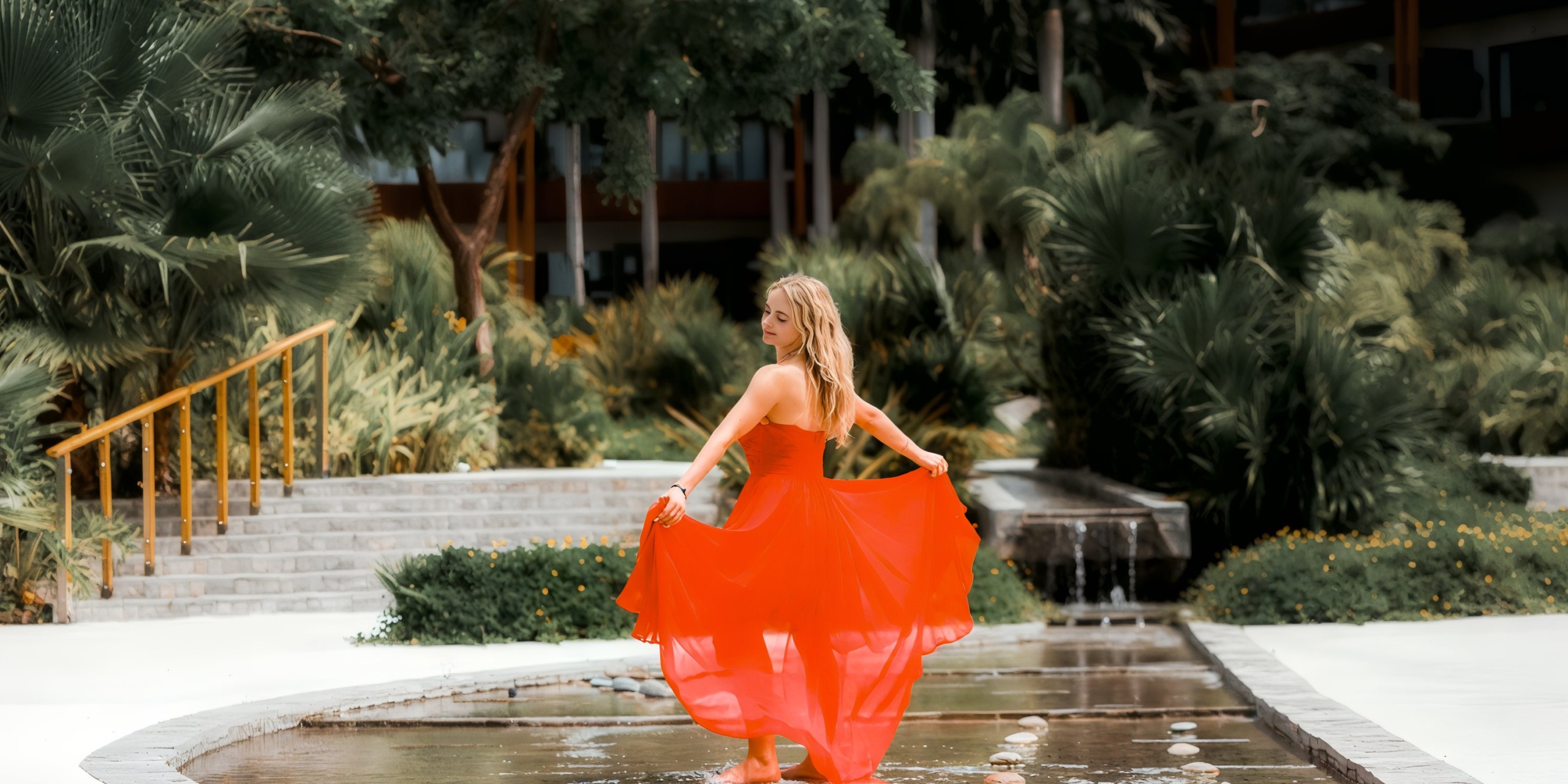 A woman wearing a dress from the Marival Armony collection stands on stepping stones in a pond, surrounded by lush greenery and trees.
