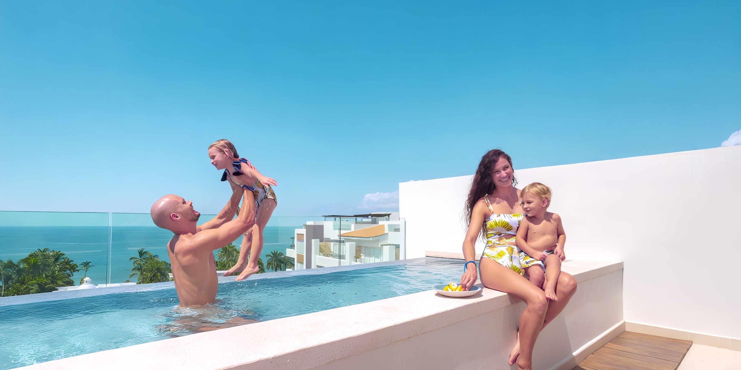 At Marival Distinct, a man plays with a child in the rooftop pool while a woman sits on the edge with another child, all enjoying the stunning view of the clear blue sky and ocean.