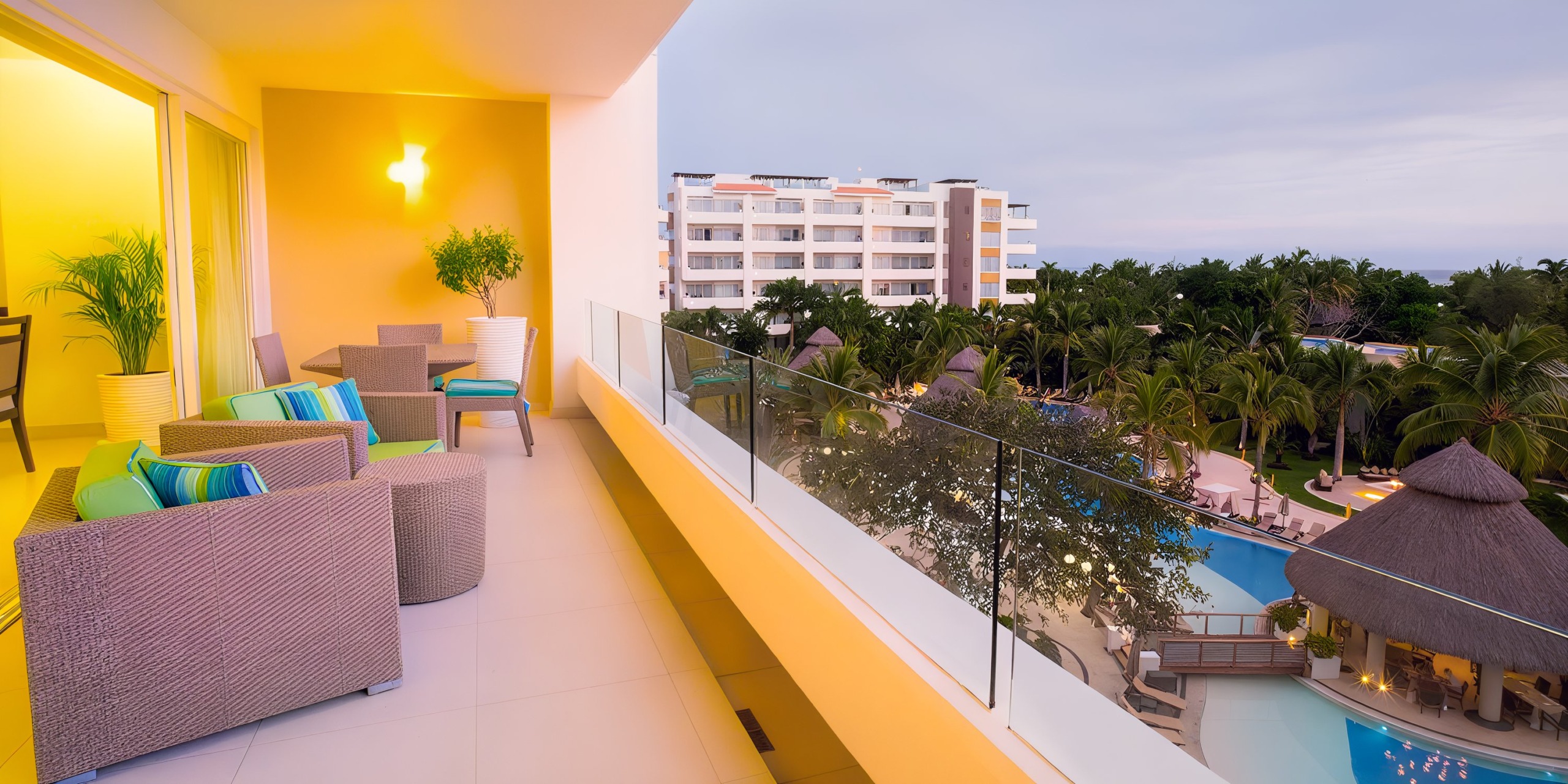 A balcony view at Marival Distinct features wicker chairs, a glass barrier, and a pool encircled by palm trees, with a multi-story building visible in the background.