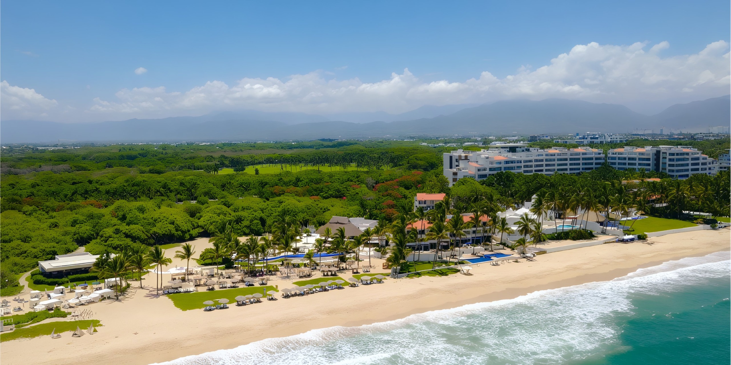 Aerial view of Marival Distinct, a resort on a sandy beach with palm trees, lounge chairs, and a pool; waves crash on the shore under a clear blue sky.