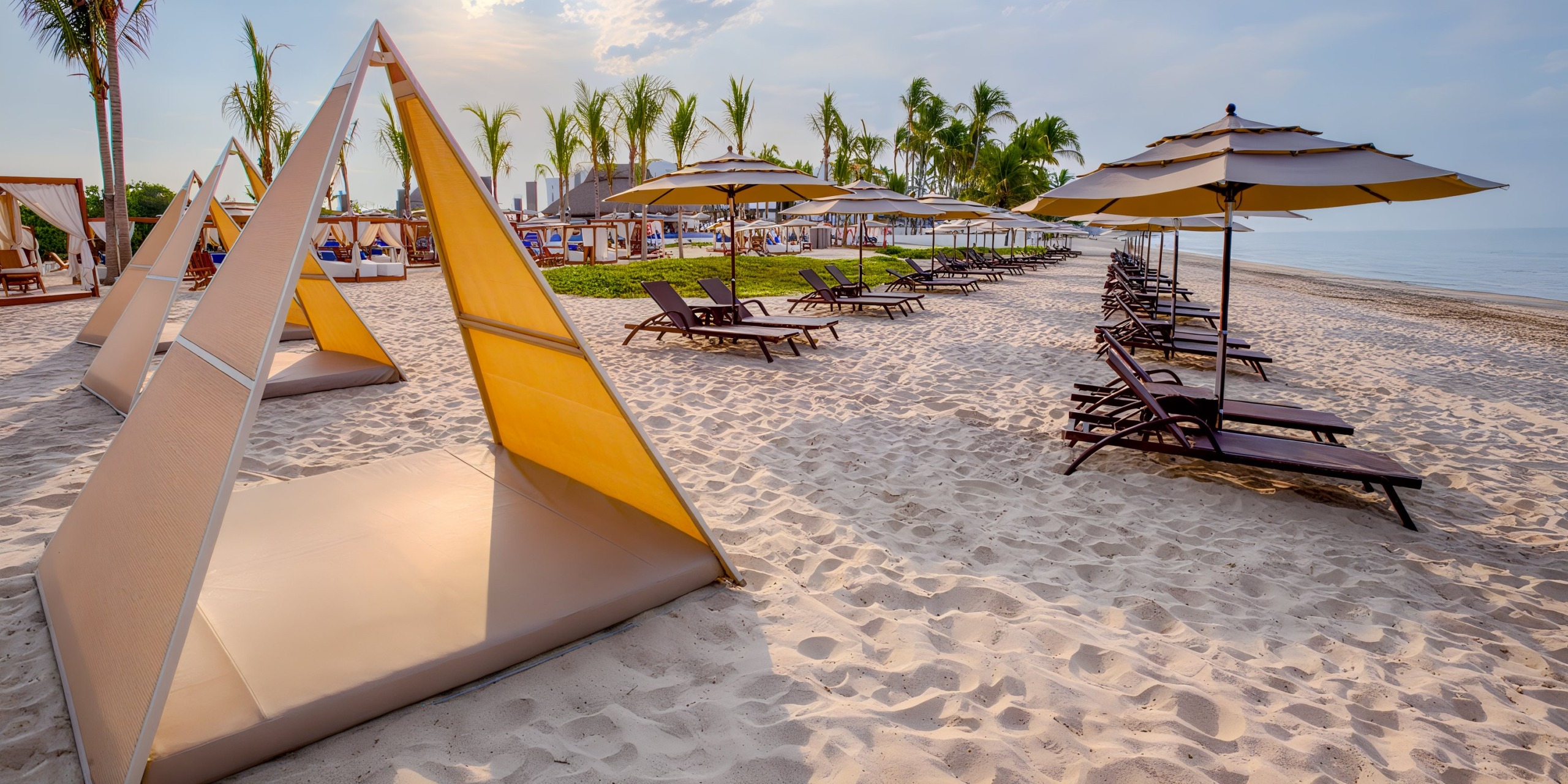 A picturesque beach featuring the Marival Distinct sun loungers, umbrellas, and triangular cabanas on pristine white sand. Tall palm trees sway gently against a backdrop of a calm sea under a clear blue sky.