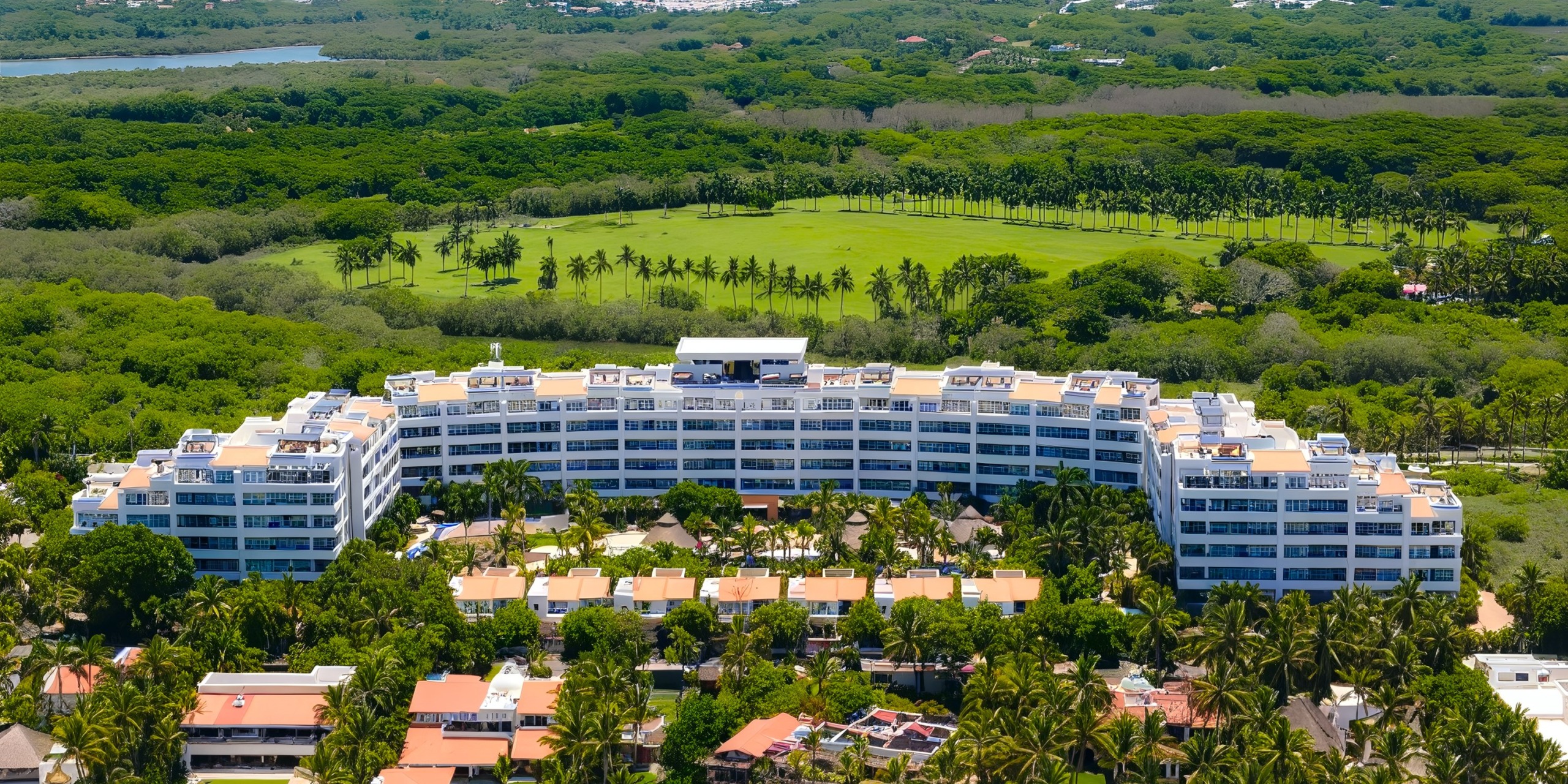 Aerial view of the expansive, curved white Marival Distinct building nestled among lush green trees and gardens, with a golf course visible in the background.