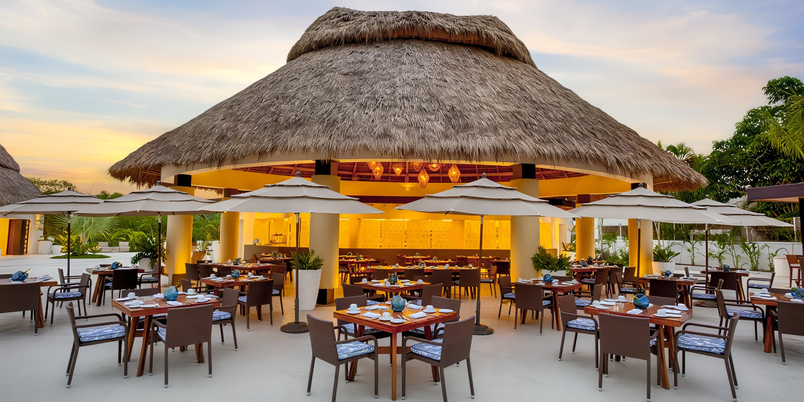 Marival Distinct features an outdoor restaurant with a charming thatched roof, complemented by wooden tables and umbrellas. The setting is beautifully surrounded by lush greenery and a clear blue sky serves as the perfect backdrop.