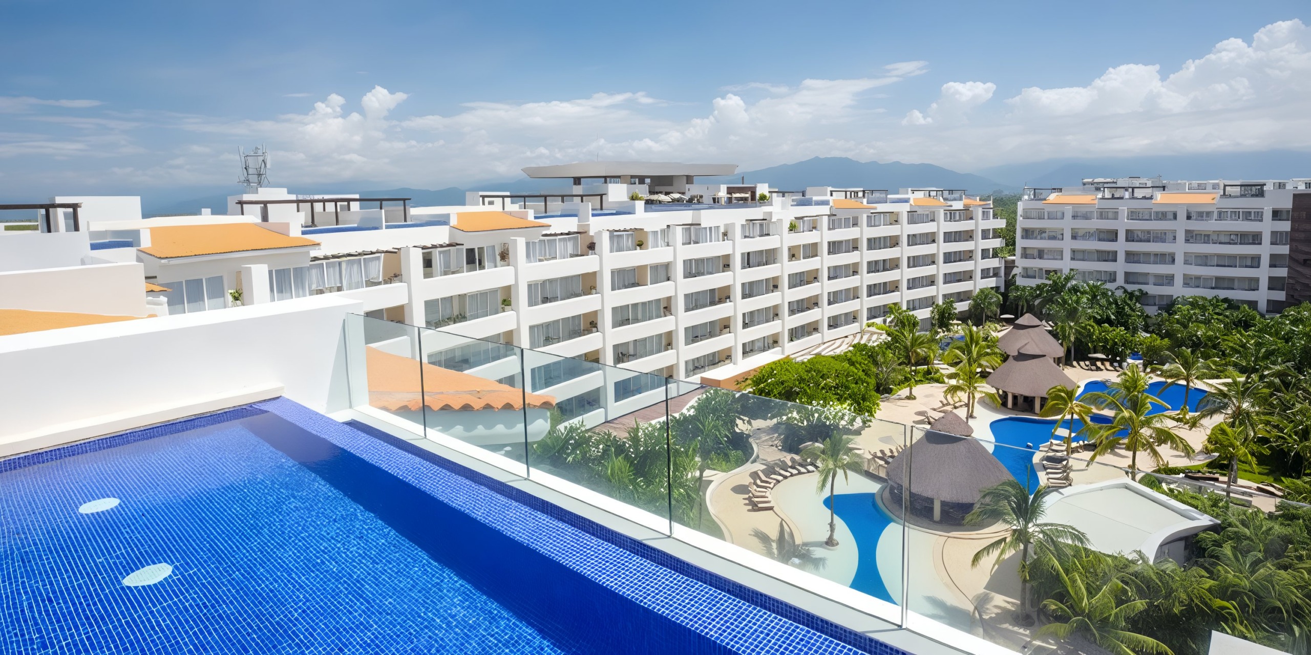 Enjoy a breathtaking view from the rooftop pool at Marival Distinct, overlooking a resort complex featuring multiple white buildings, beautifully landscaped gardens, and two winding blue pools beneath a partly cloudy sky.