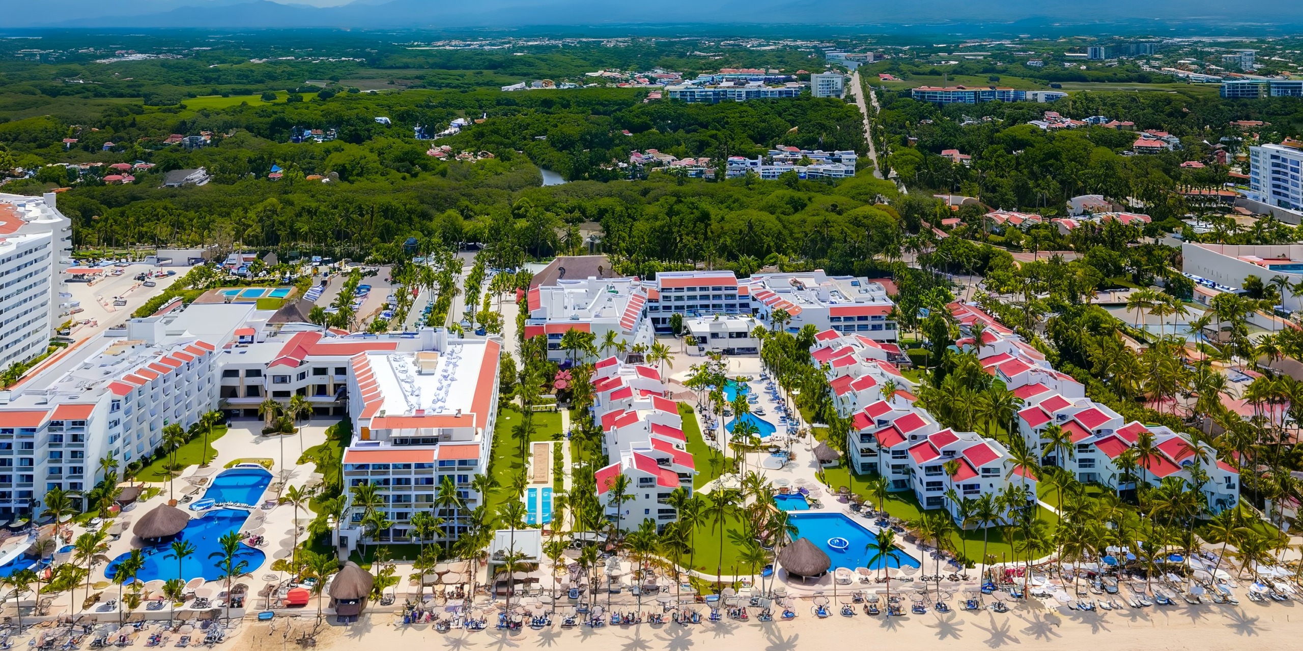 Aerial view of Marival Emotions, a beachfront resort featuring red-roofed buildings, swimming pools, and palm trees, all set amidst lush greenery with distant mountain views.