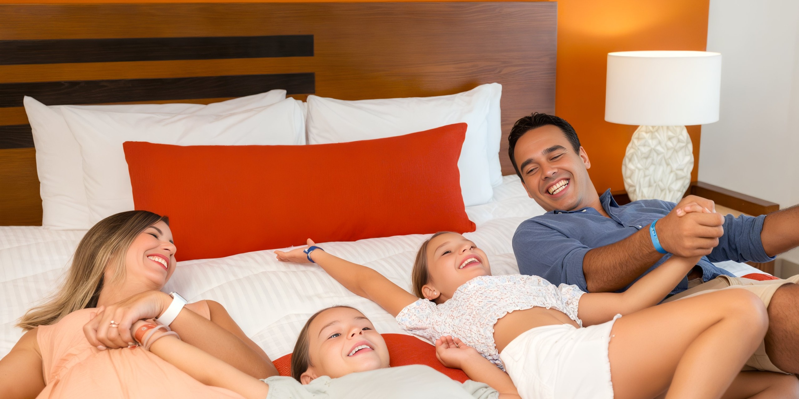A family of four laughing and relaxing on a bed adorned with Marival Emotions orange and white pillows in a warmly lit bedroom.