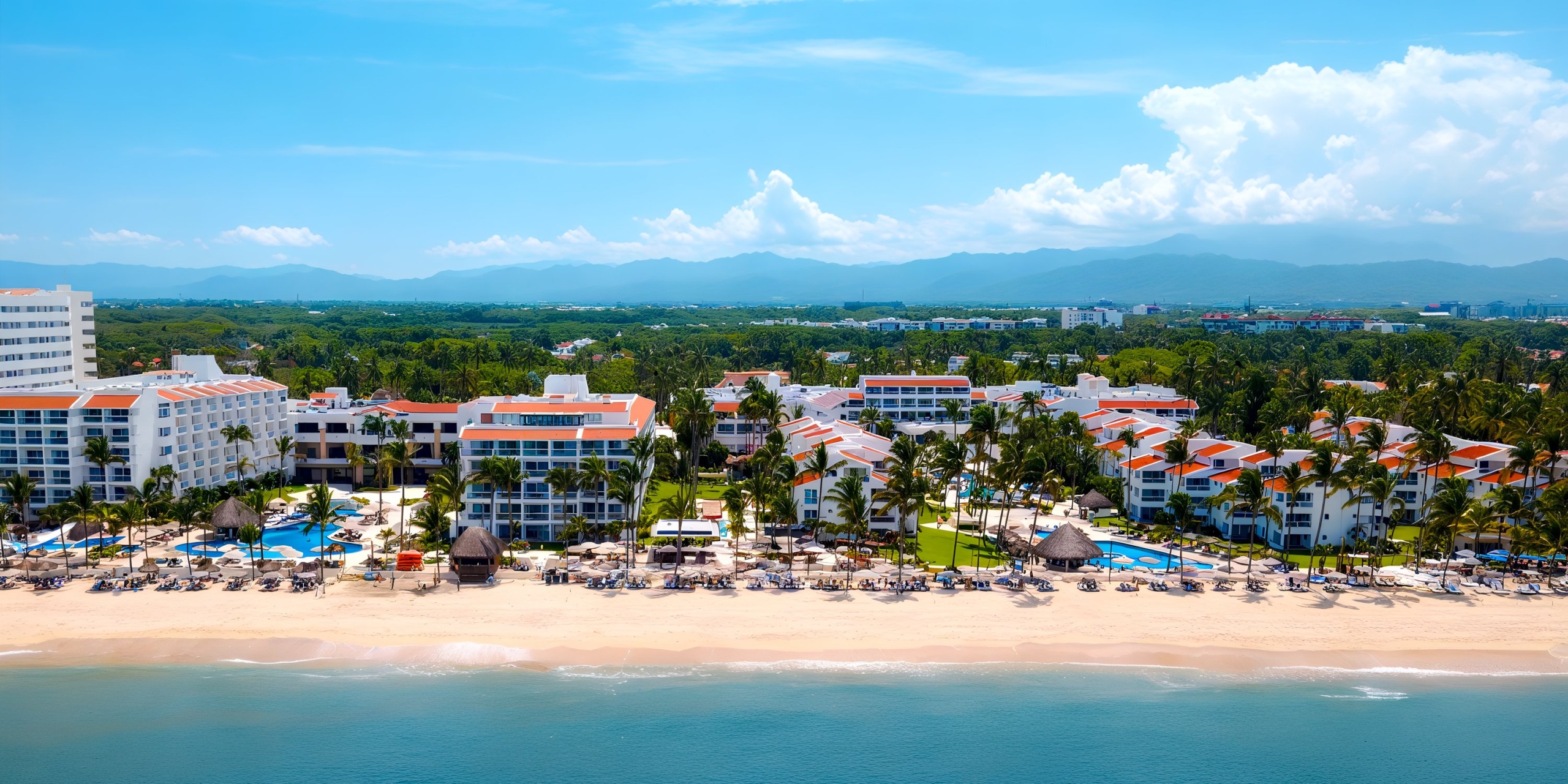 Experience the breathtaking aerial view of Marival Emotions, a beachfront resort featuring red-roofed buildings, surrounded by palm trees and lounge chairs that dot the sandy shore. In the background, lush green hills and clouds complete this picturesque setting.