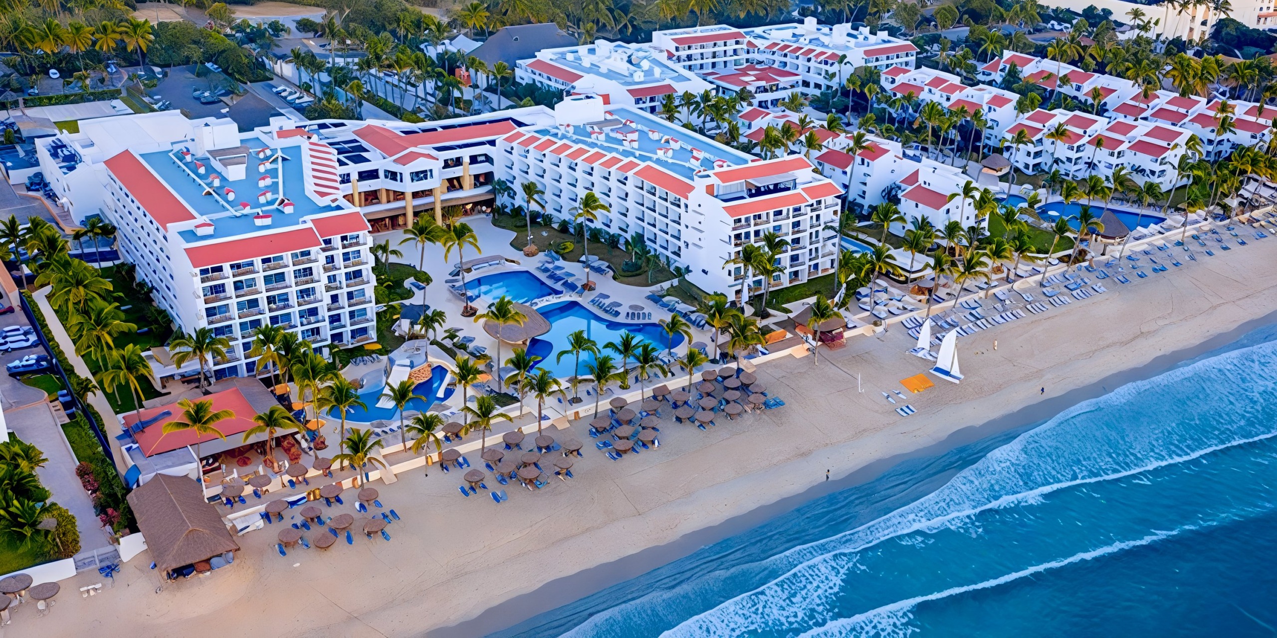 Aerial view of Marival Emotions, a beachfront resort featuring multiple white buildings, swimming pools, palm trees, and beach chairs facing the ocean.