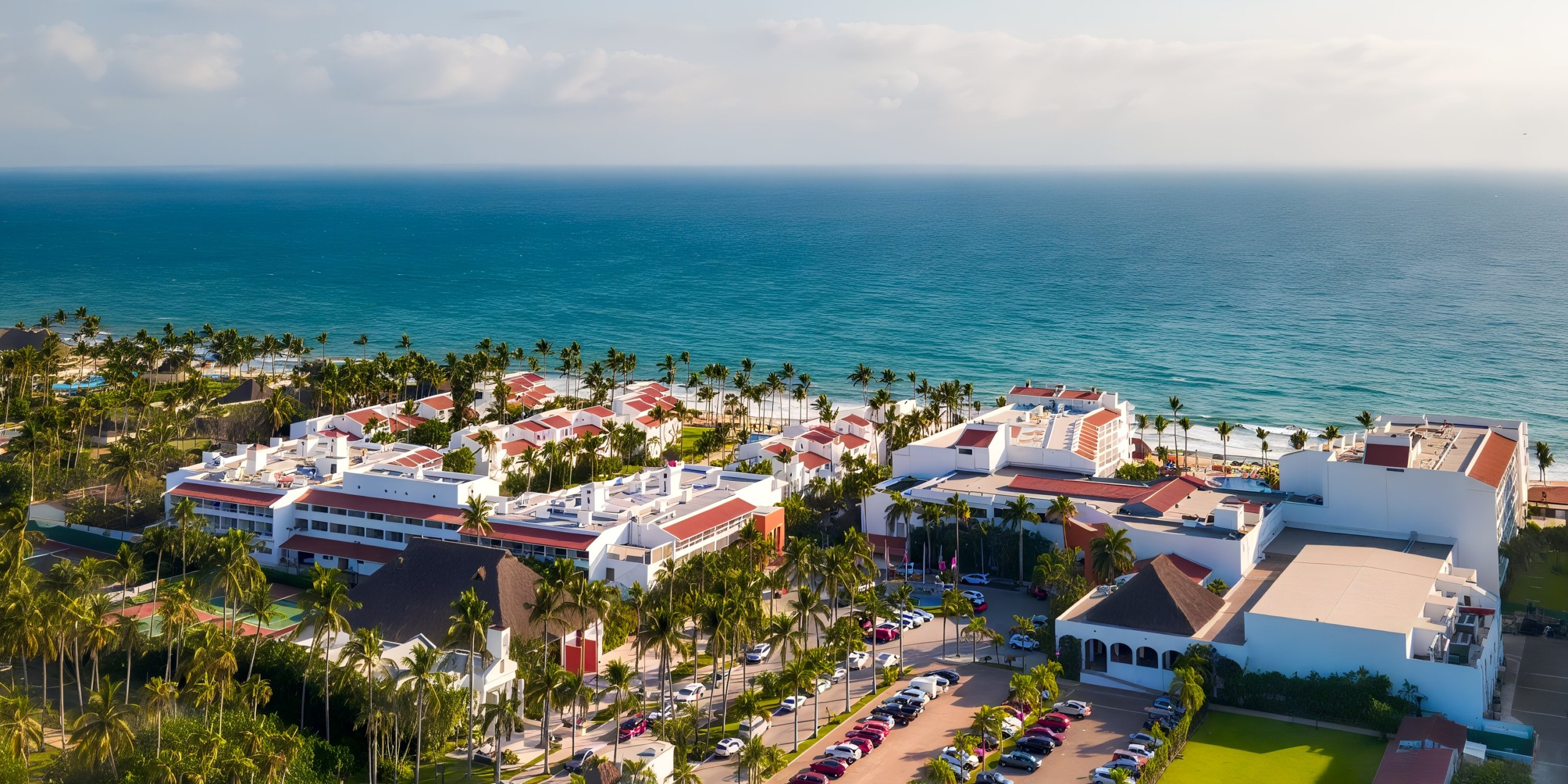 Aerial view of Marival Emotions, an oceanfront resort featuring red-roofed buildings surrounded by palm trees and parking lots, adjacent to a blue sea under a partly cloudy sky.