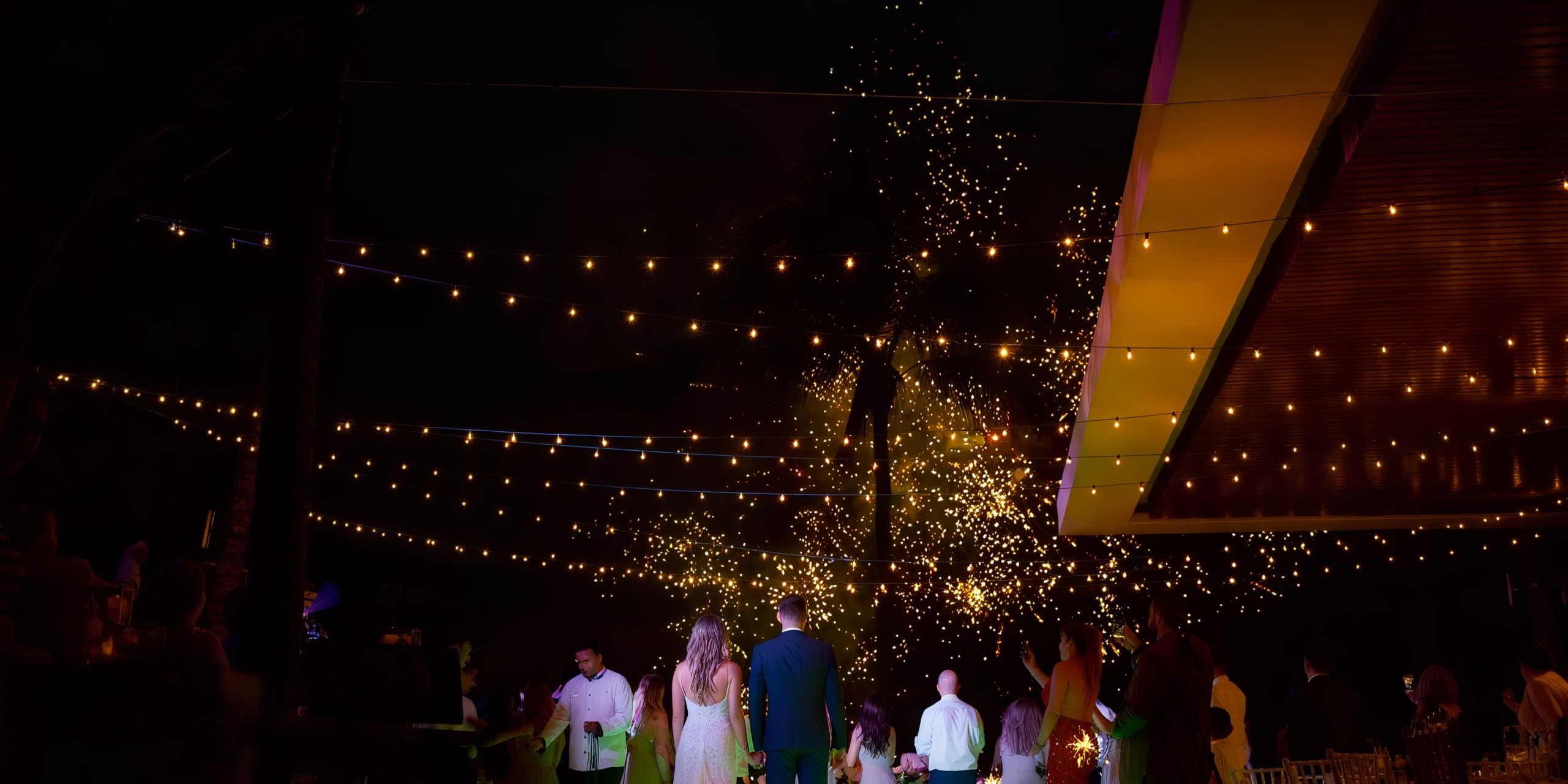 A group of people gathered under the Marival Emotions string lights, watching fireworks against a dark night sky.