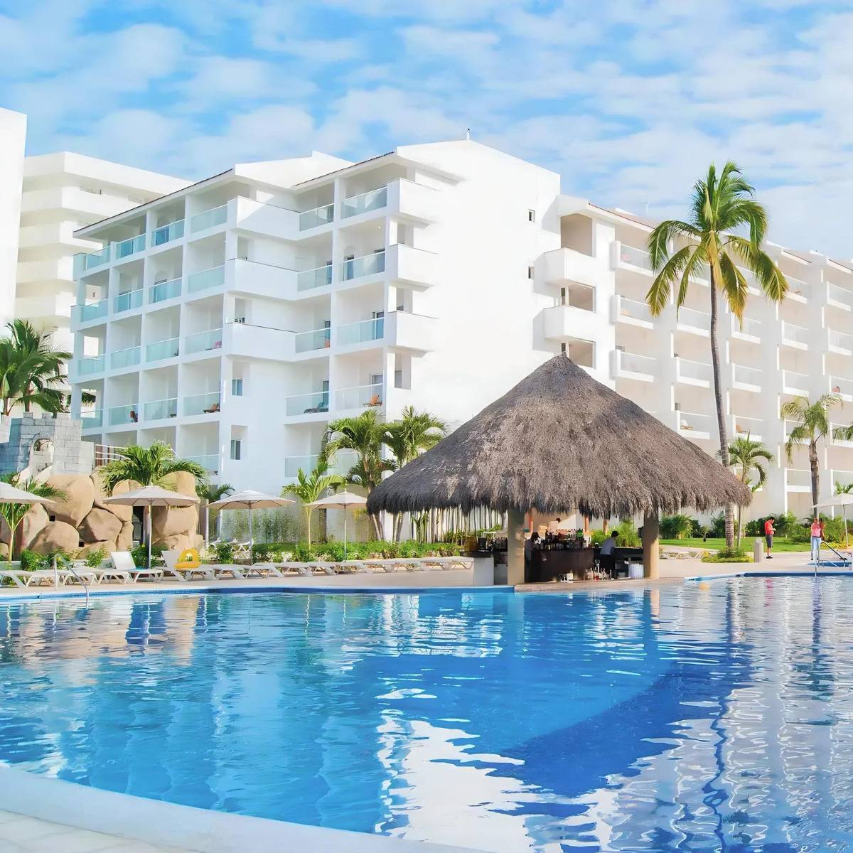 Modern white Marival Emotions resort building with a thatched poolside bar and a clear blue swimming pool, perfect for a destination wedding, located under a bright blue sky.