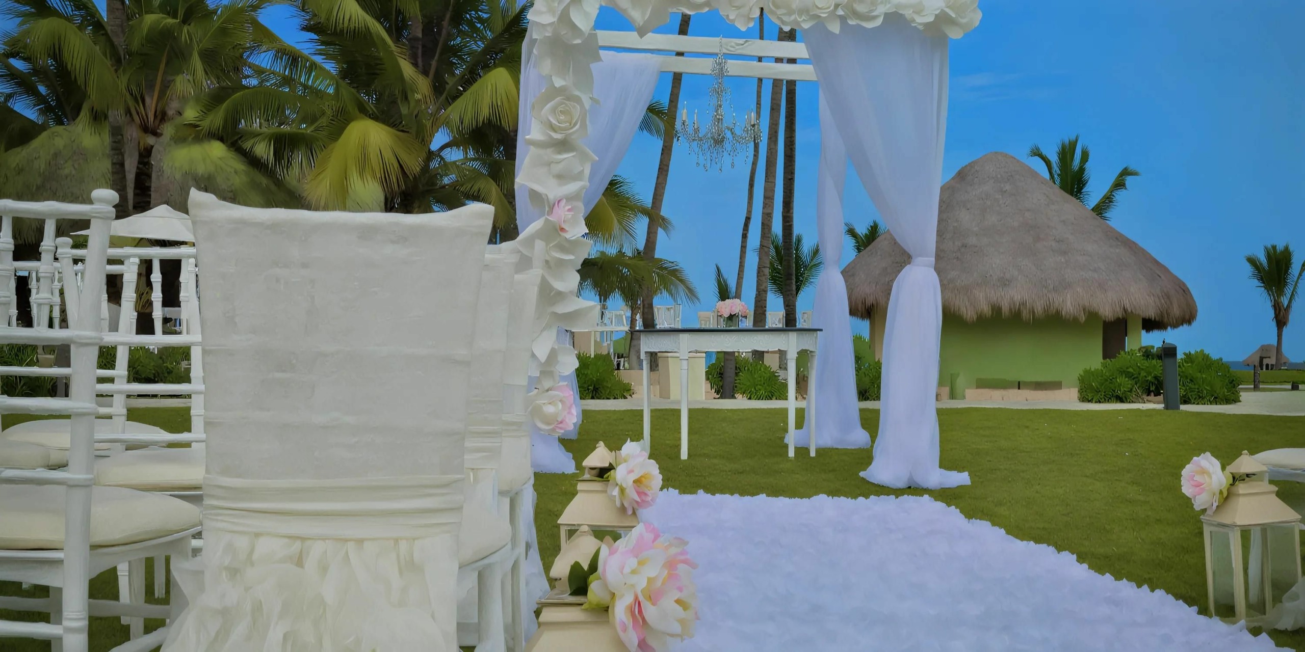 Dreams Onyx: An outdoor wedding setup featuring pristine white chairs, a beautifully decorated arch, elegant floral arrangements, and a luxurious white carpet aisle on a lush grassy area adjacent to majestic palm trees and a charming thatched-roof building.