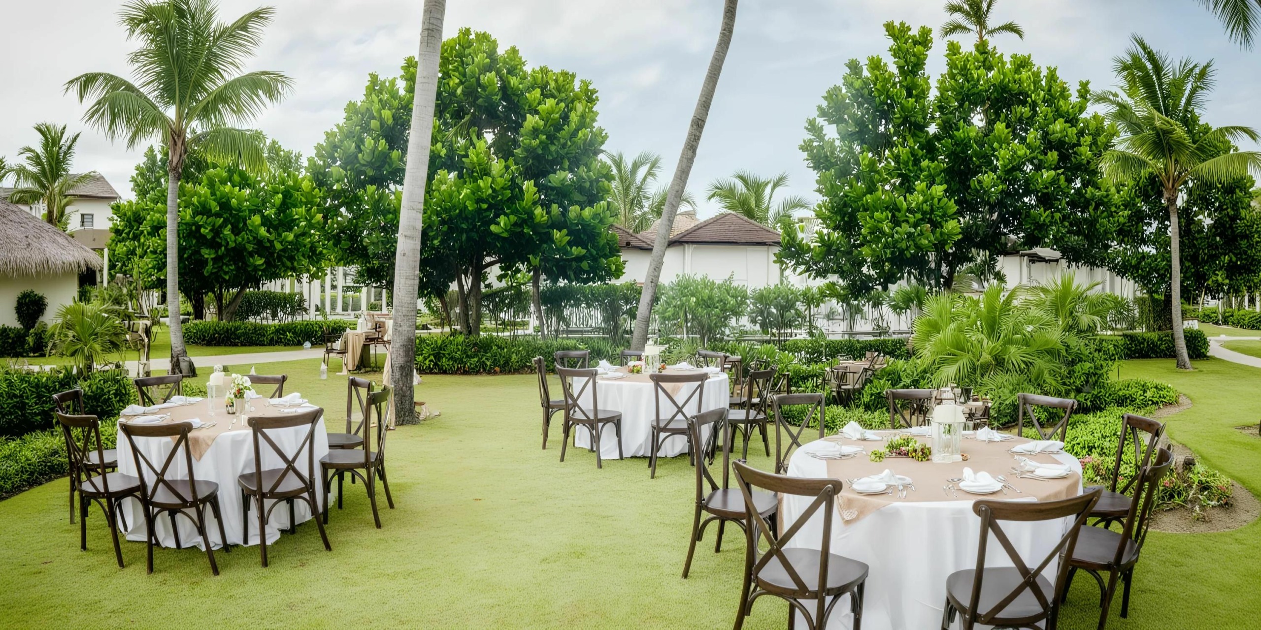 Outdoor dining setup with several Dreams Onyx round tables covered with white tablecloths and wooden chairs, located on a grassy area surrounded by palm trees and lush greenery.