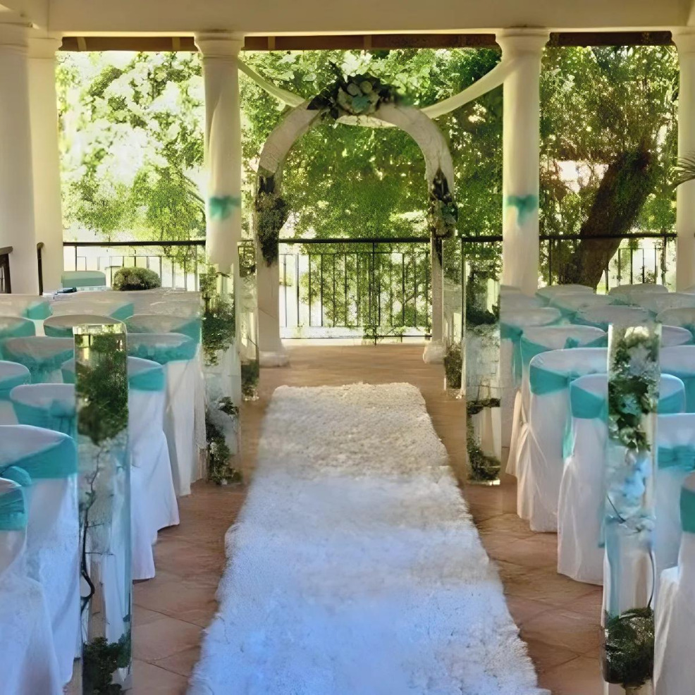 A destination wedding setup with a white aisle runner lined by chairs with teal sashes under a columned gazebo, overlooking greenery.