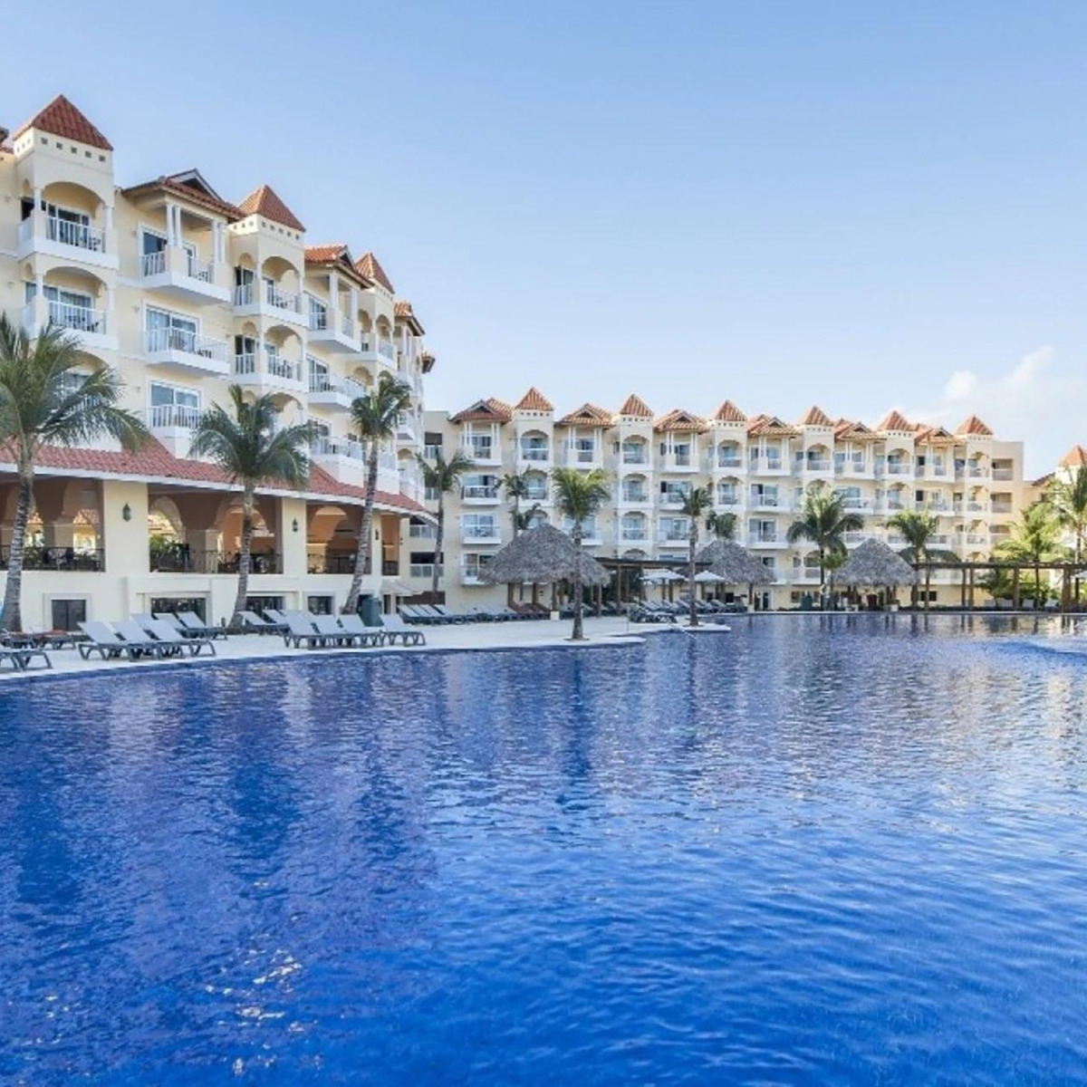 A popular Occidental Caribe destination weddings venue featuring a large swimming pool with clear blue water in front of a multi-story hotel with red-tiled roofs under a clear sky.