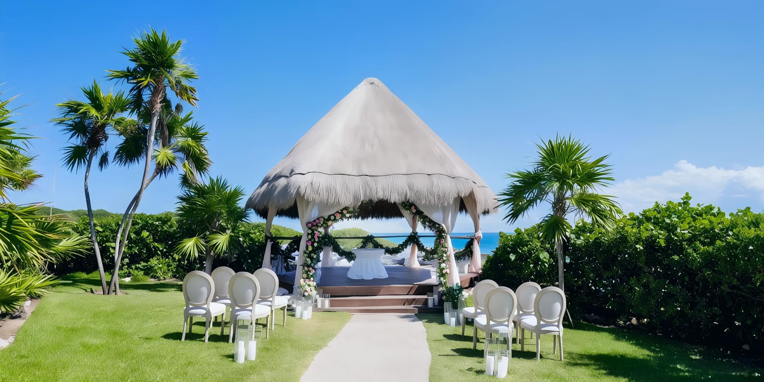A small, round, thatched-roof gazebo set up for a wedding ceremony with white chairs and floral decorations, surrounded by palm trees and greenery, near a bright blue ocean under a clear sky.