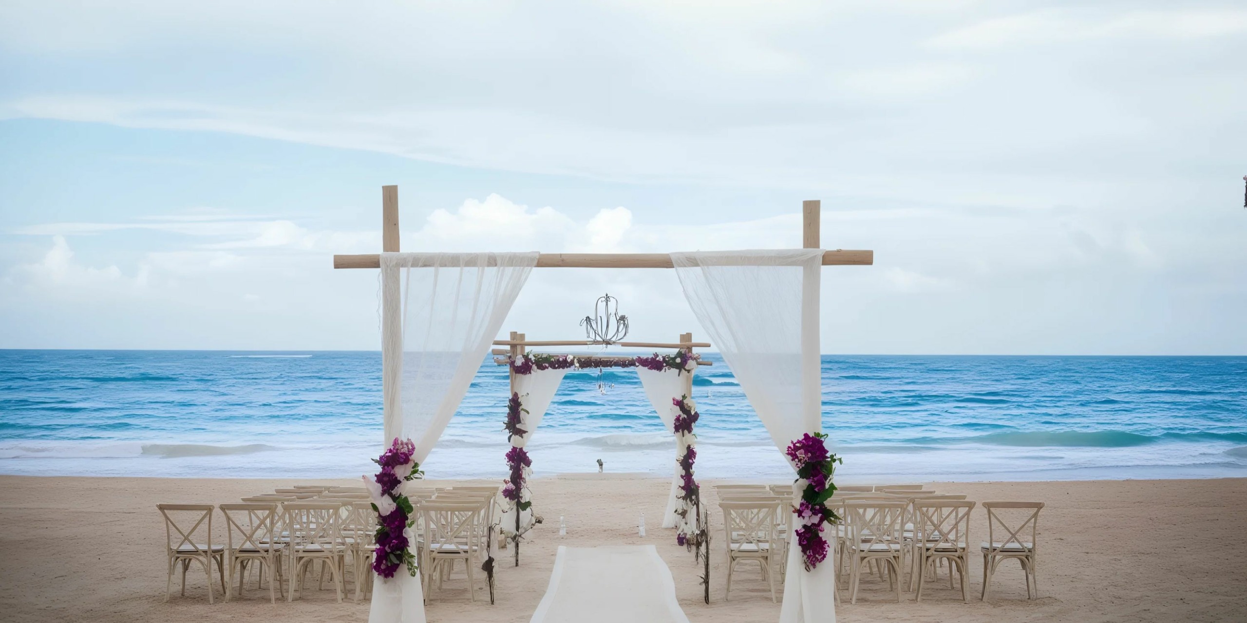 Beach wedding setup with a wooden arch decorated with white drapes and purple flowers, facing the ocean. Rows of chairs are arranged on the sand.