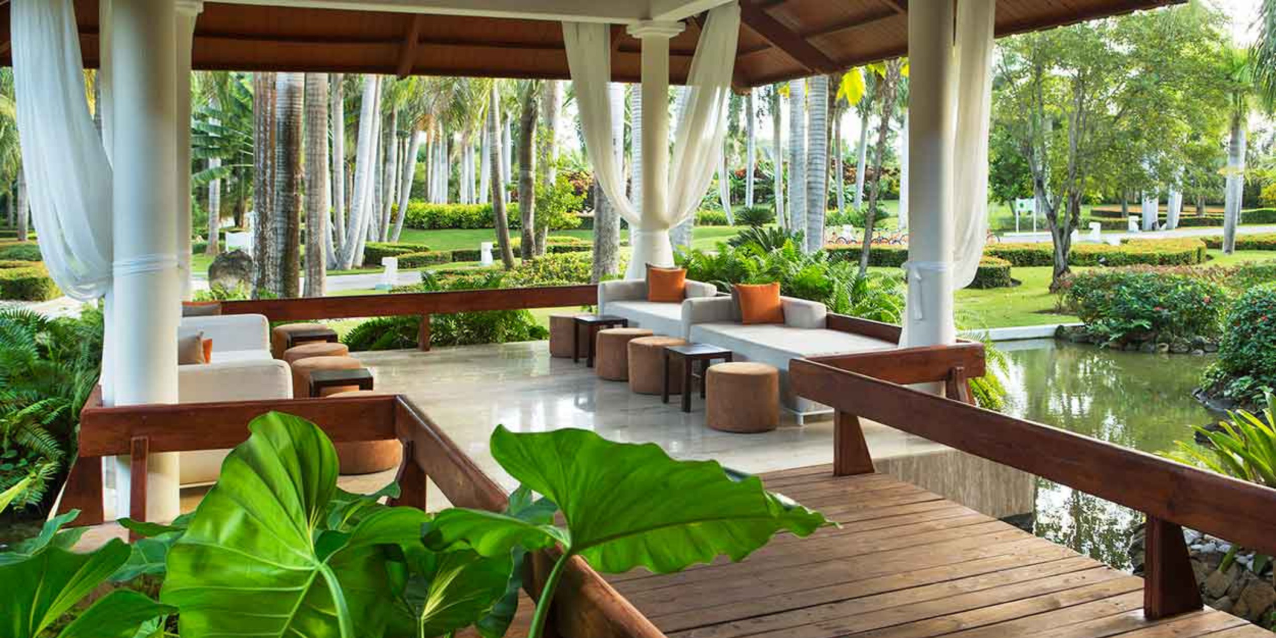 Outdoor lounge area with sofas and pillows under a pavilion, ideal for destination weddings, surrounded by lush greenery and a small pond.