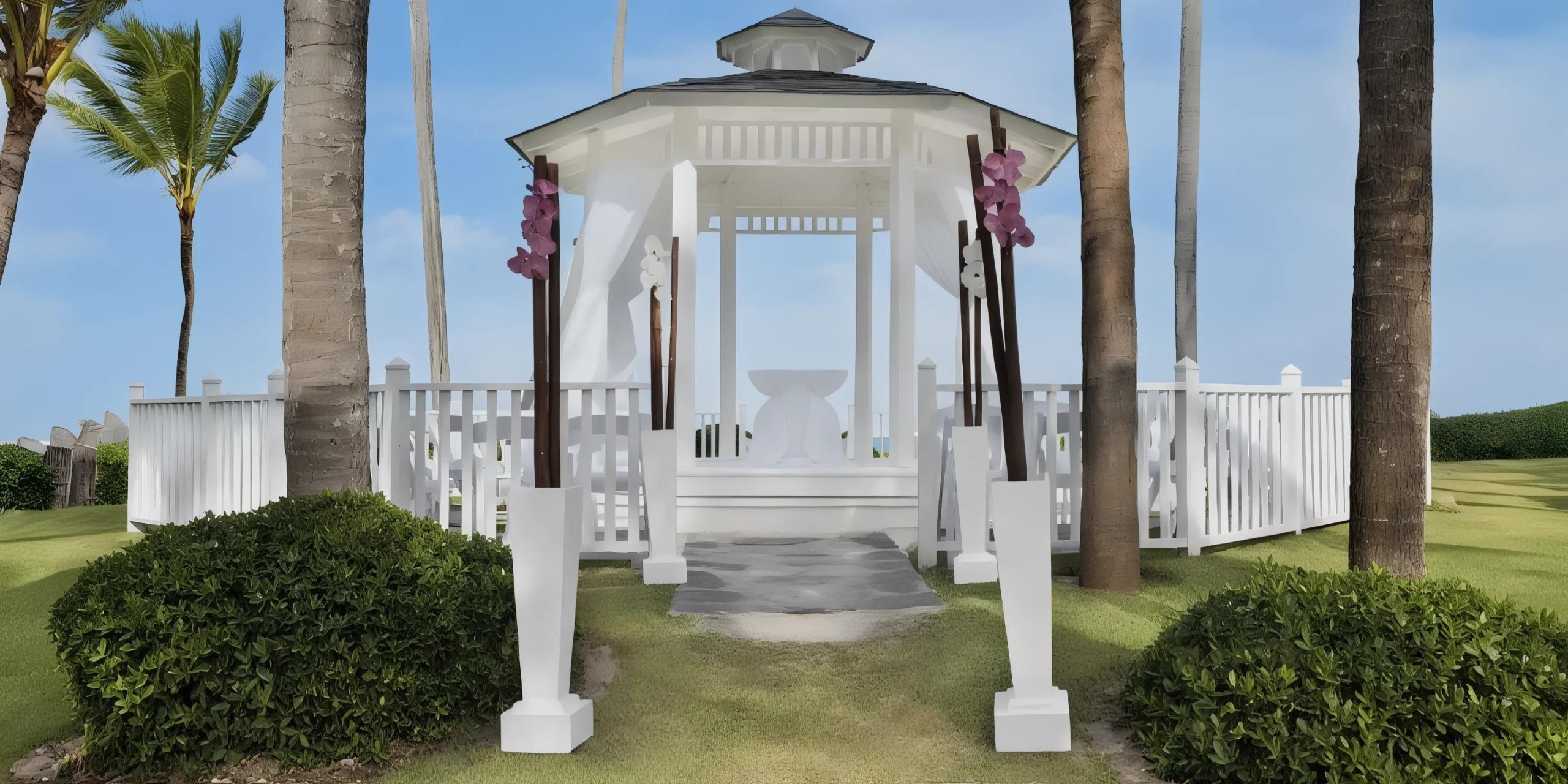 A white gazebo with a pathway, surrounded by palm trees, greenery, and white fence, set against a clear blue sky.