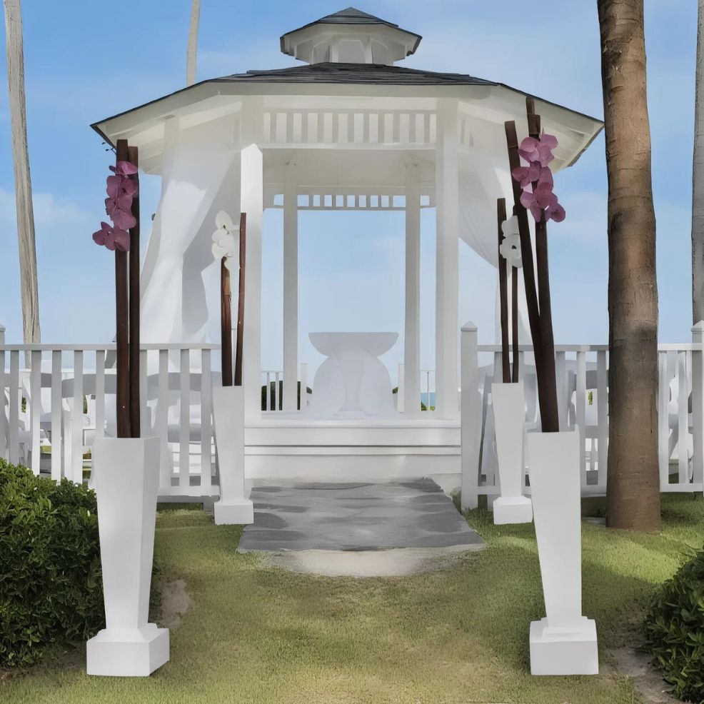 A white gazebo with a dark roof is decorated with tall vases containing pink flowers. It is situated on a grassy area, with a white picket fence and a clear sky in the background.