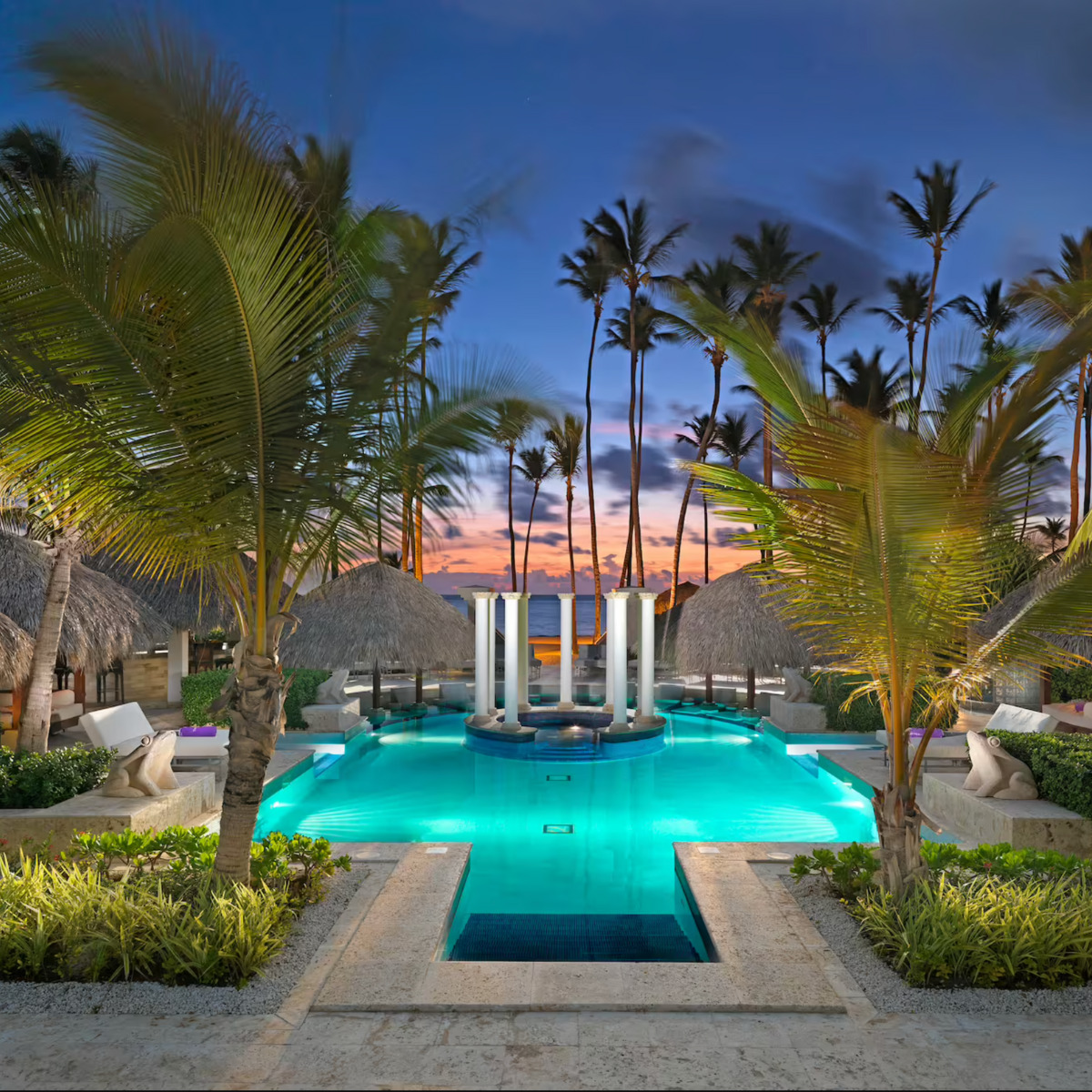 A luxurious Paradisus Punta Cana destination resort pool with illuminated water, surrounded by palm trees and thatched-roof cabanas, under a twilight sky.